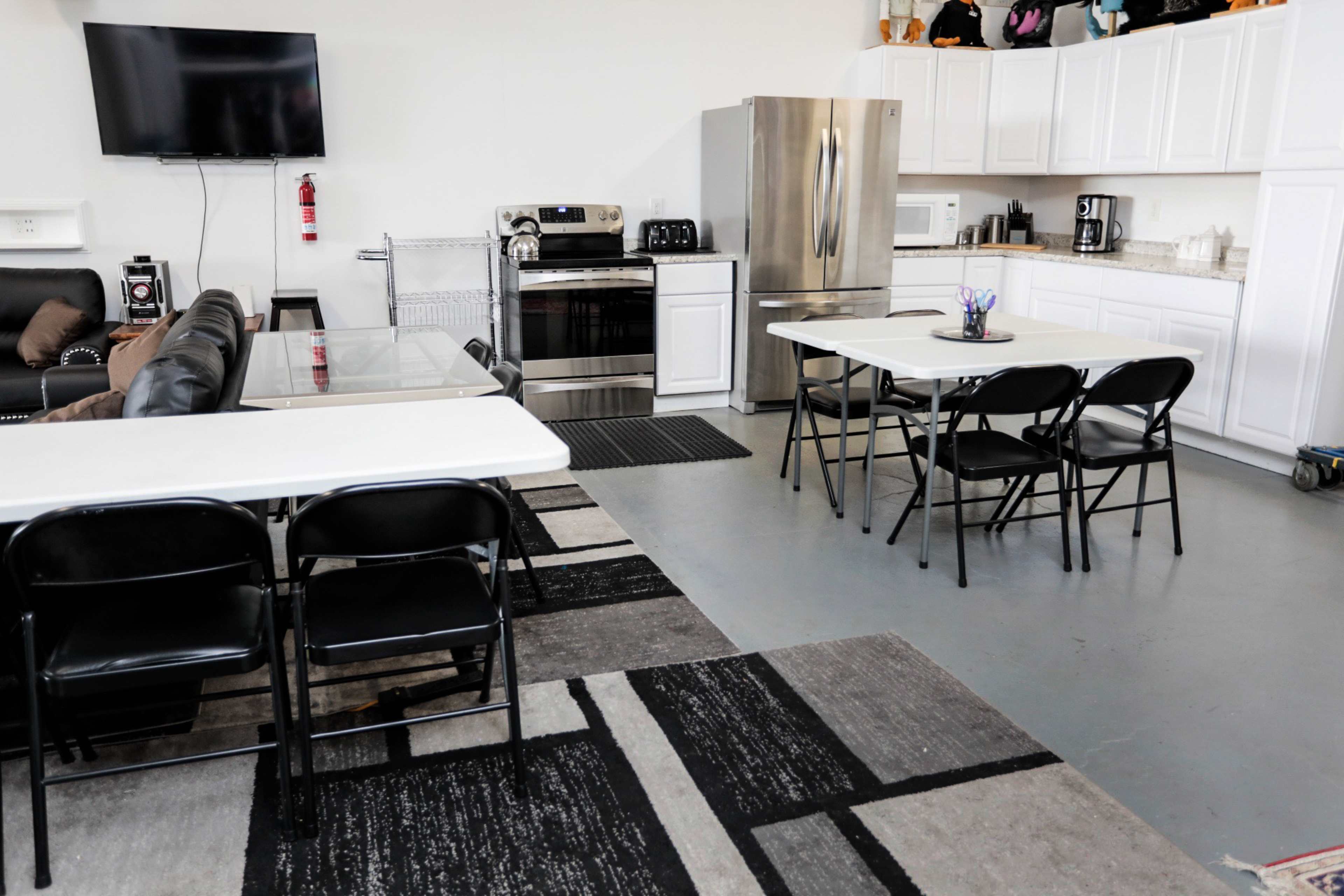 The image shows a functional kitchen and dining area featuring a large stainless steel fridge, an oven, white cabinets, a black couch, and several tables with chairs.