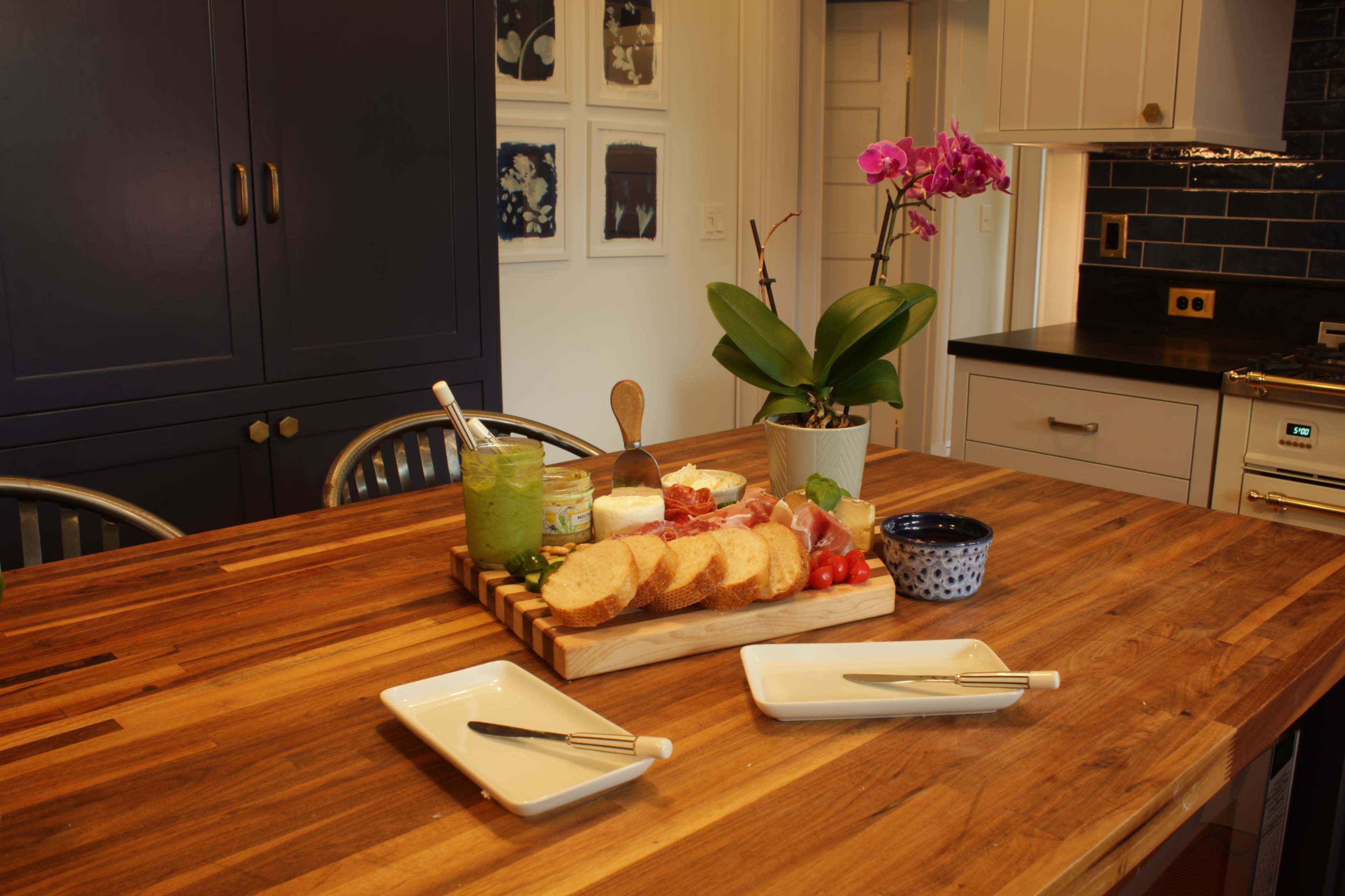 A wooden serving board on a kitchen island displays an assortment of sliced bread, dips, and garnishes, accompanied by two empty plates and utensils.