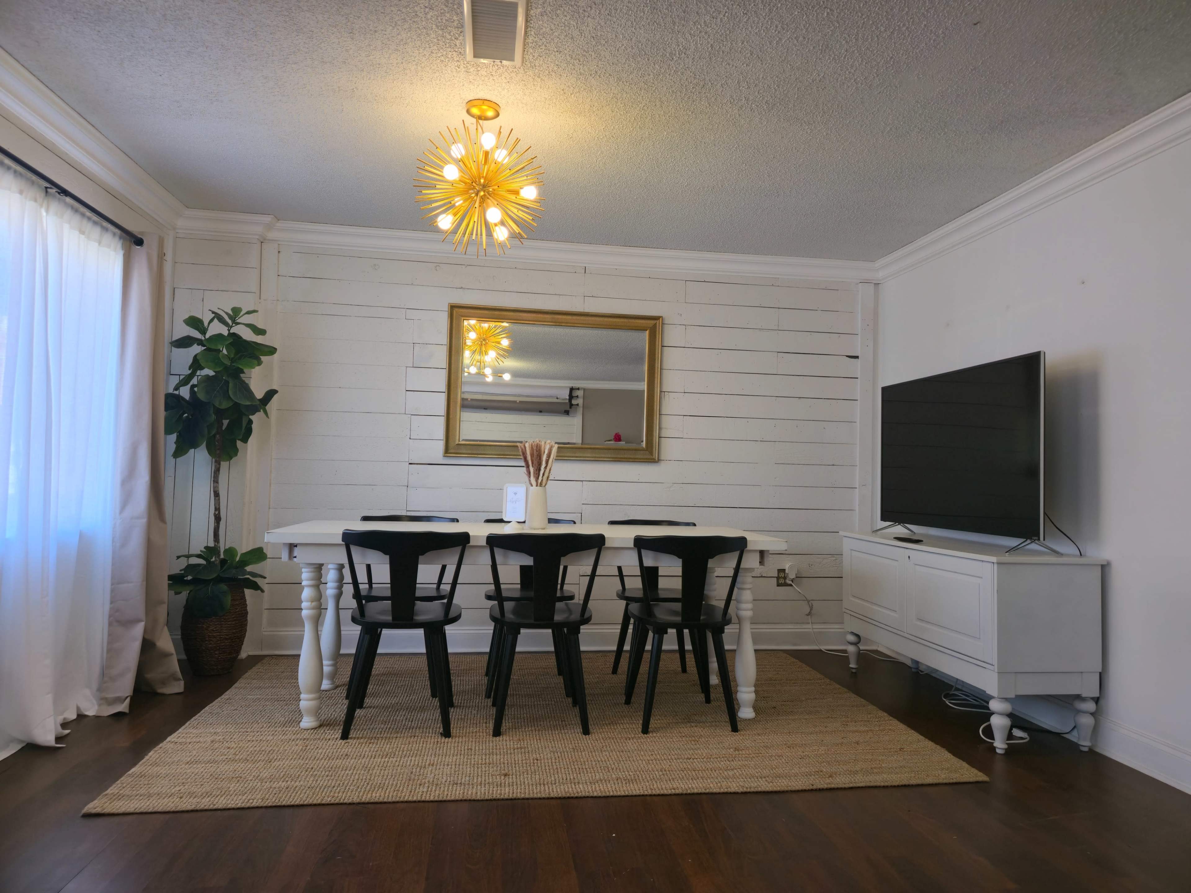 The image shows a dining area featuring a round white table surrounded by black chairs, a large television mounted on the wall, a mirror with a gold frame, and a decorative plant in a corner.