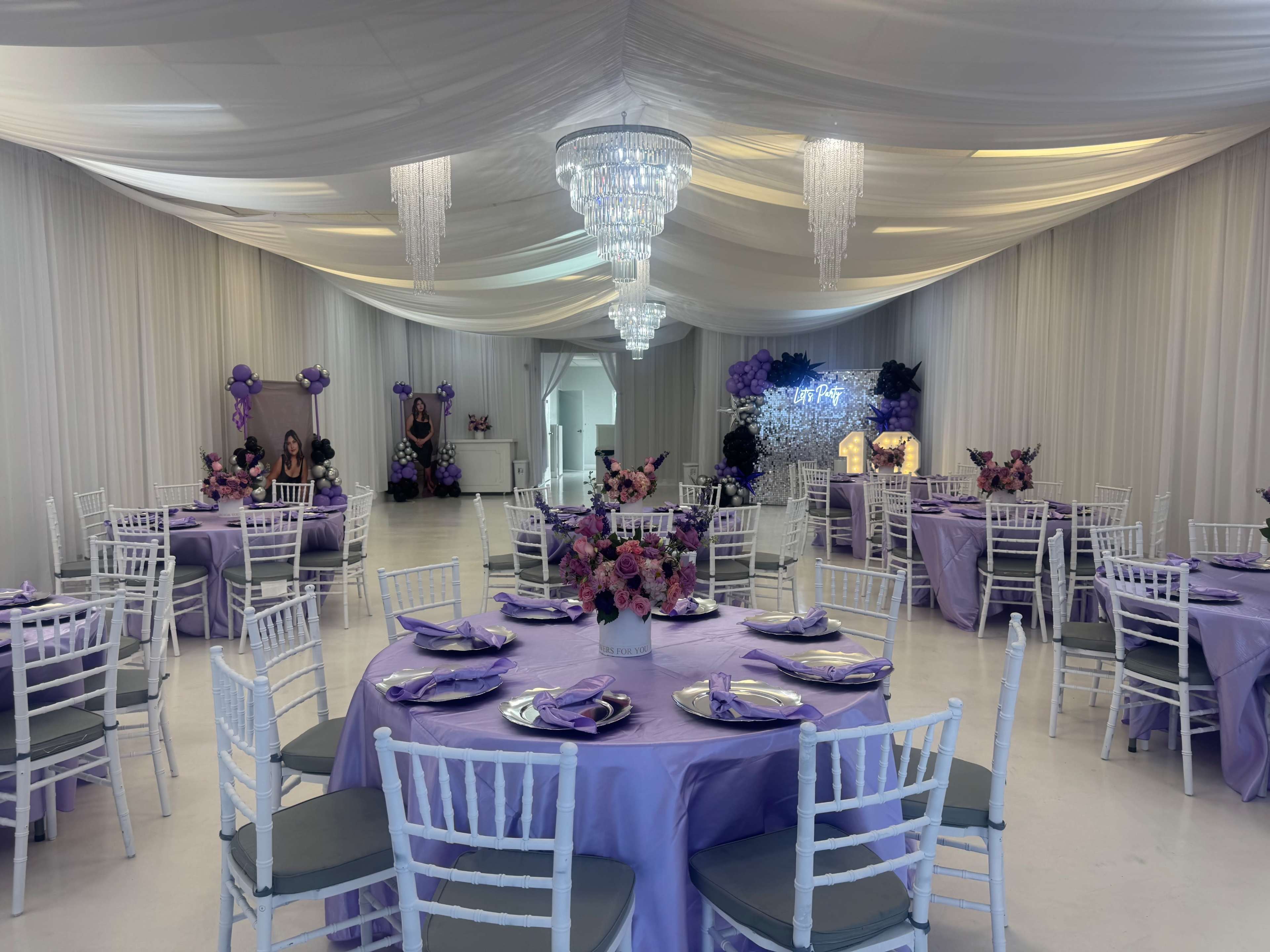 The image shows an elegantly decorated event space with round tables covered in purple tablecloths and adorned with floral centerpieces, all beneath draped white fabric and hanging chandeliers.