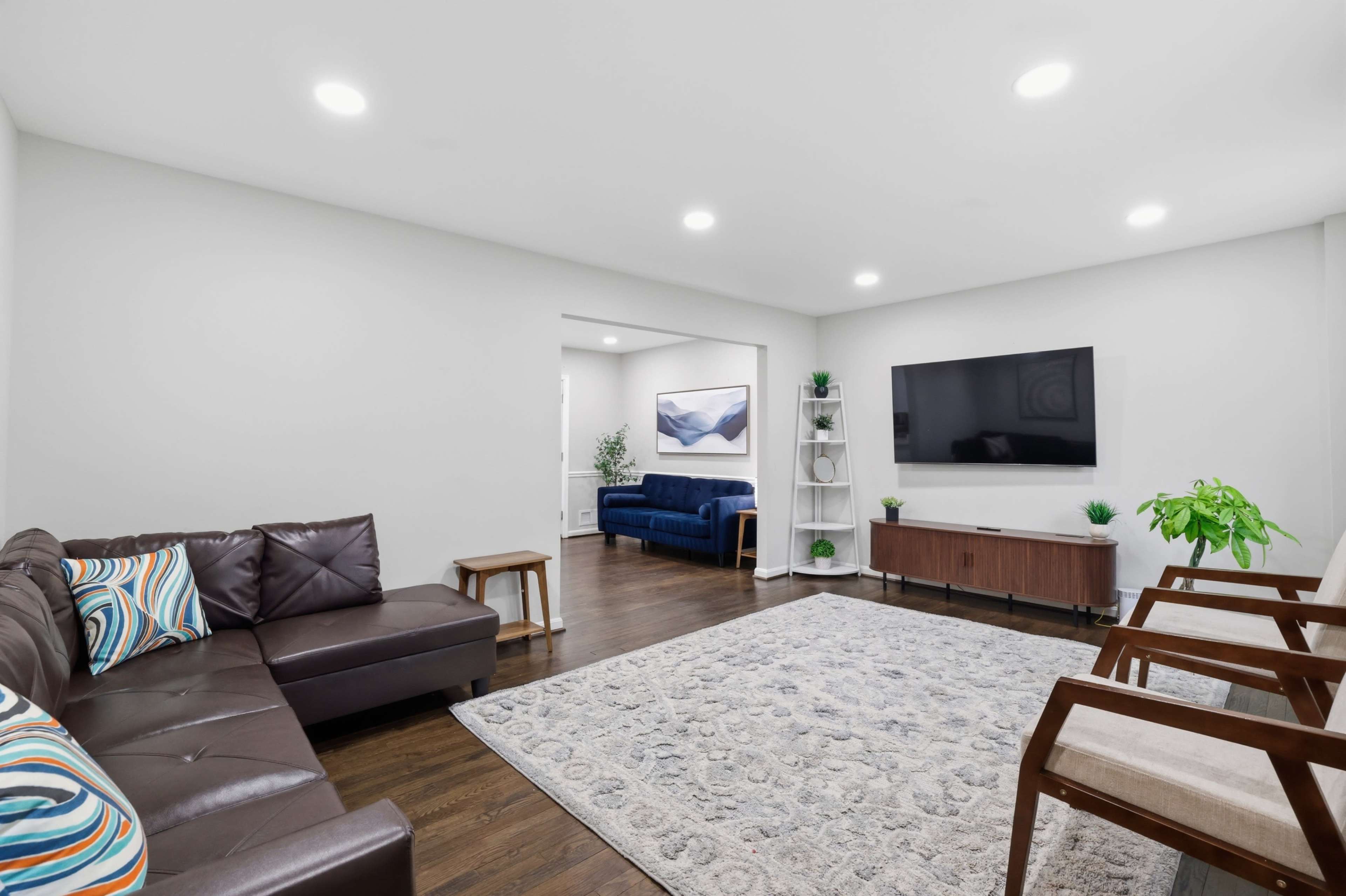 A spacious living room features a leather sectional sofa, a blue couch, a large wall-mounted TV, and a rug with a decorative pattern.