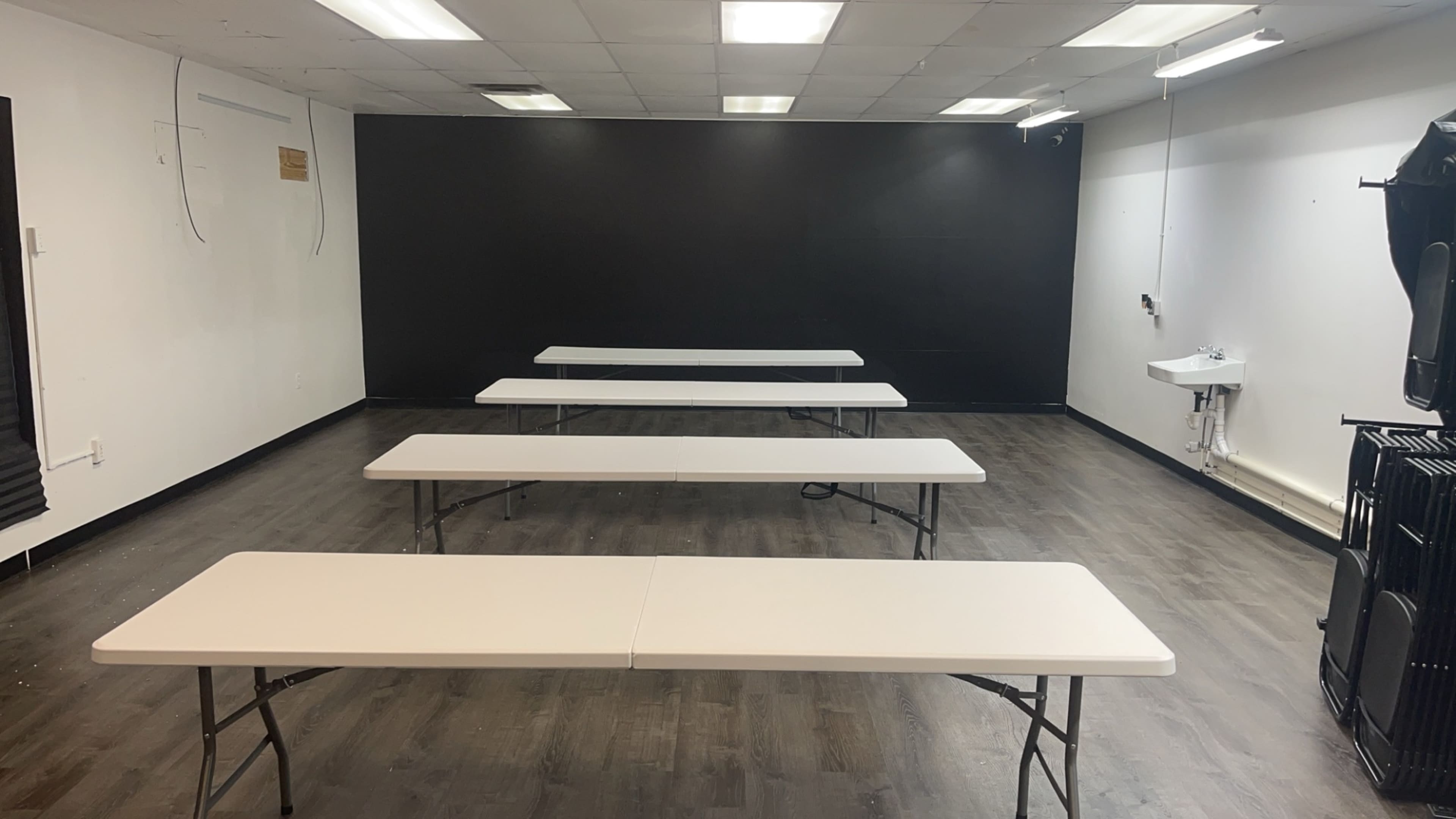 A spacious room with five rectangular tables arranged in a line, a black wall in the background, and a sink along one side.