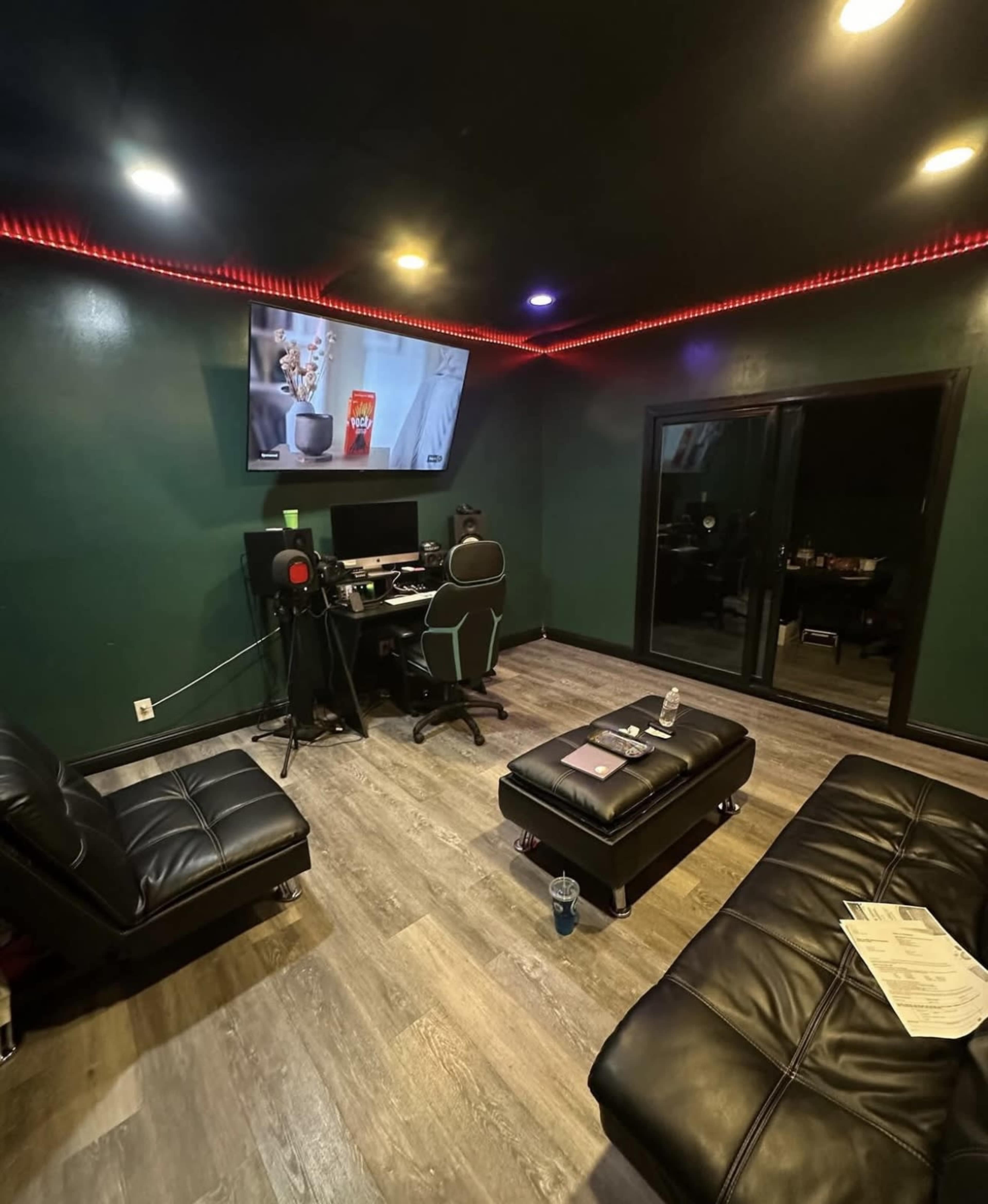 The image shows a studio room with a dark green wall, a desk with computer equipment, a large TV mounted on the wall, and two black sofas arranged around a small table.
