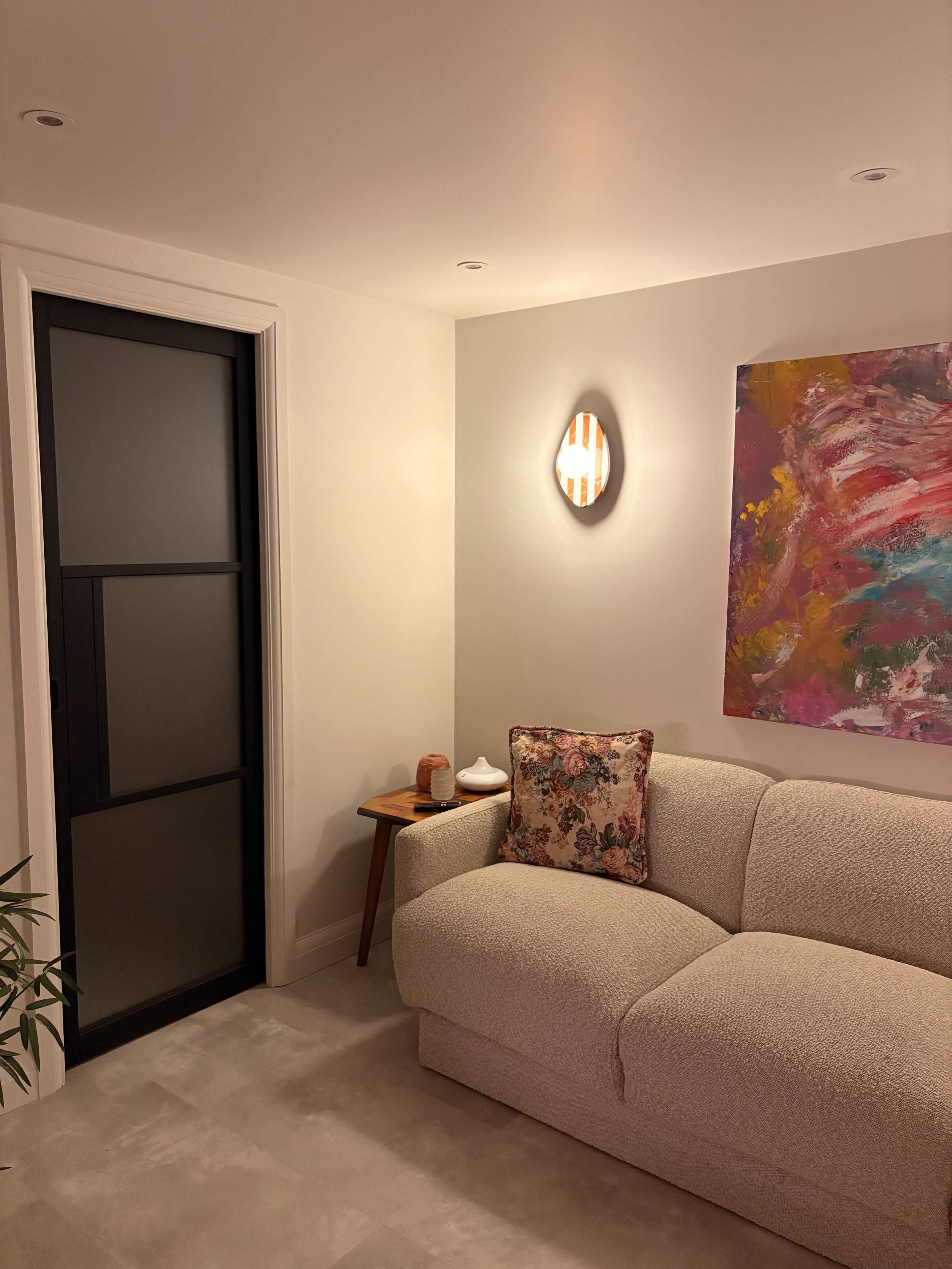 The image shows a cozy living room with a textured white sofa, a small wooden side table, a colorful abstract painting on the wall, and a doorway with a glass panel.