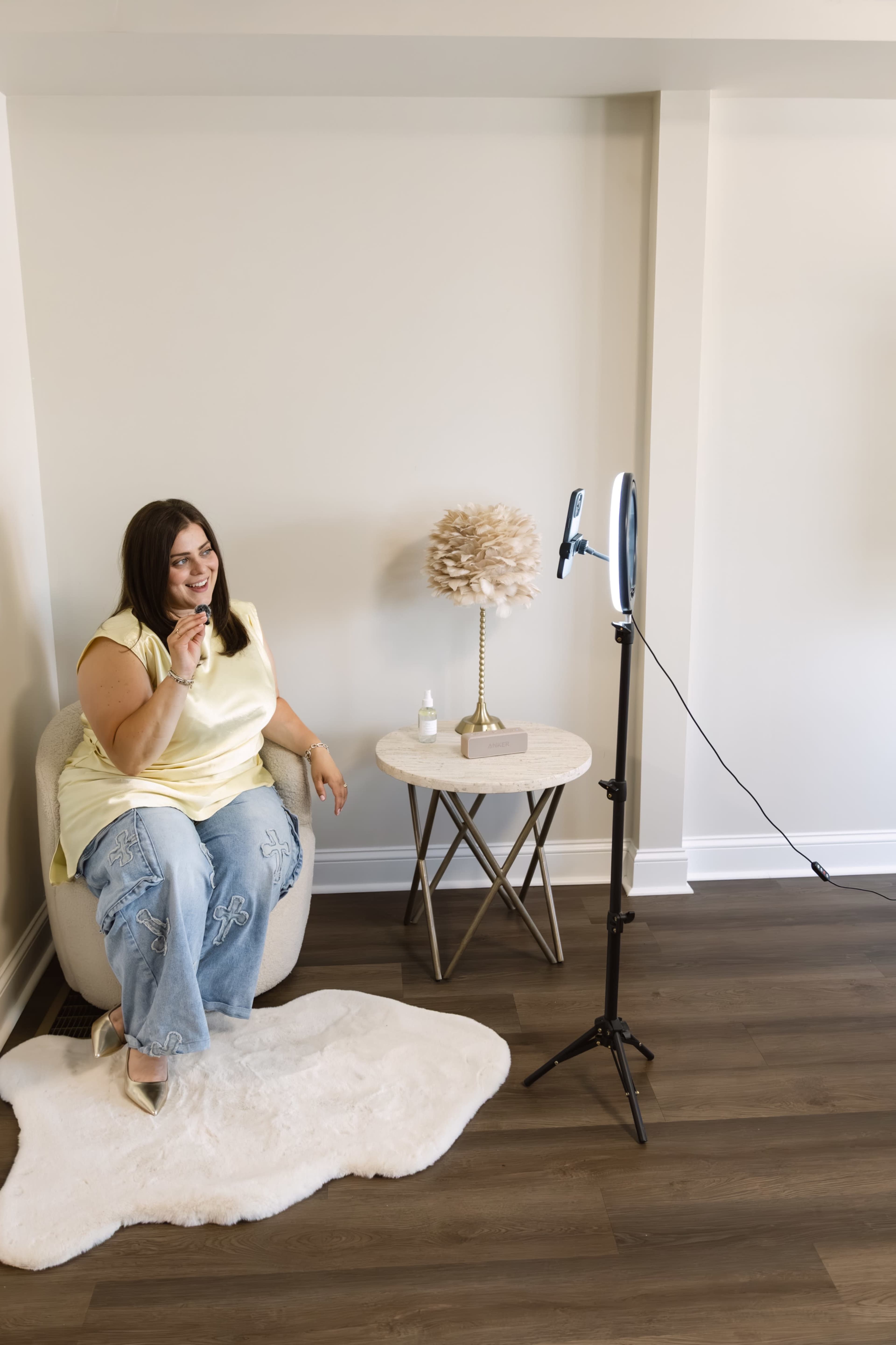 A woman sits on a chair in a bright room, smiling and gesturing toward a ring light positioned on a tripod next to her.