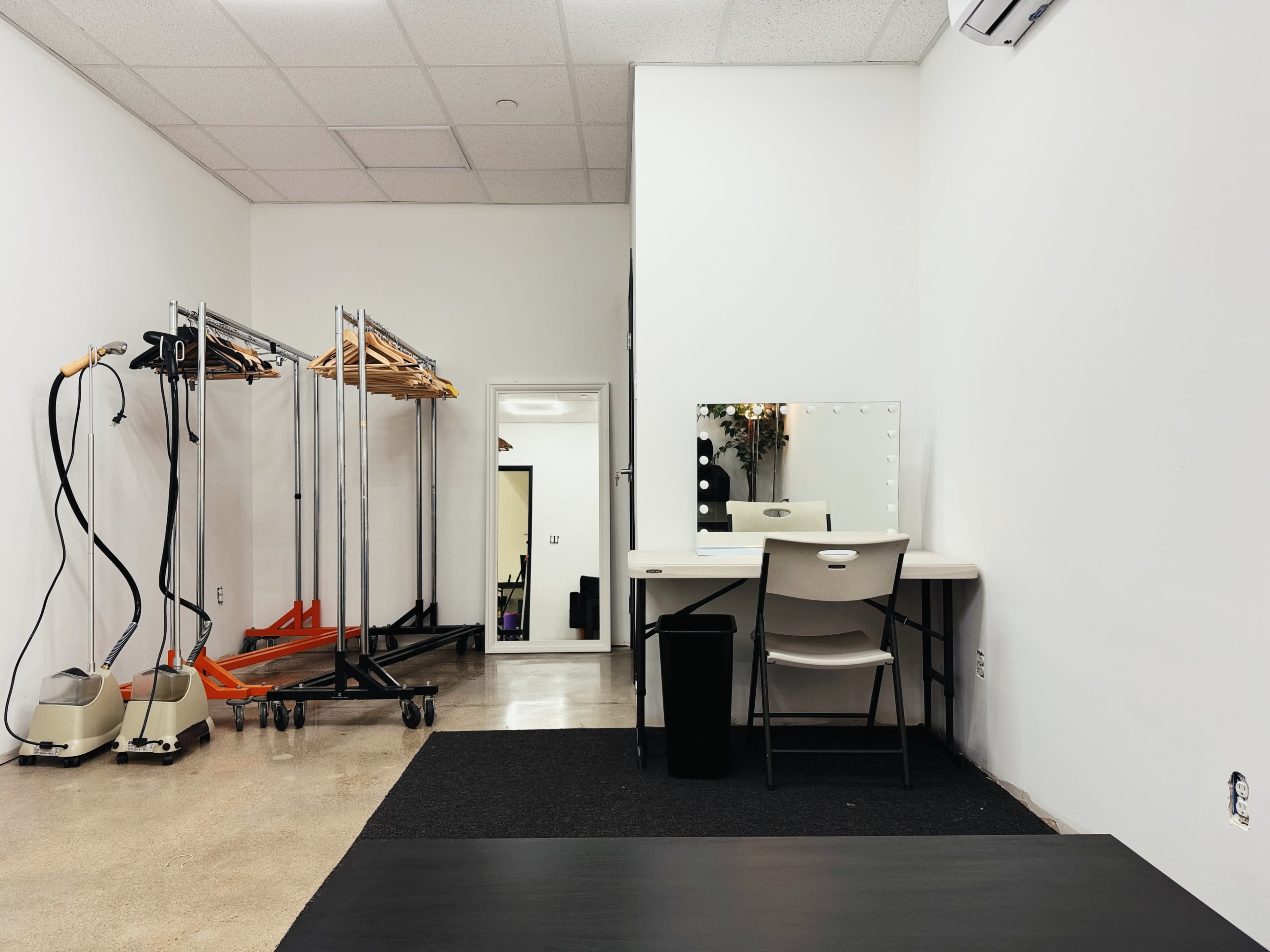 The image shows a minimalist dressing room with a mirror and chair on a table, racks for clothing, and a small black wastebasket in the corner.