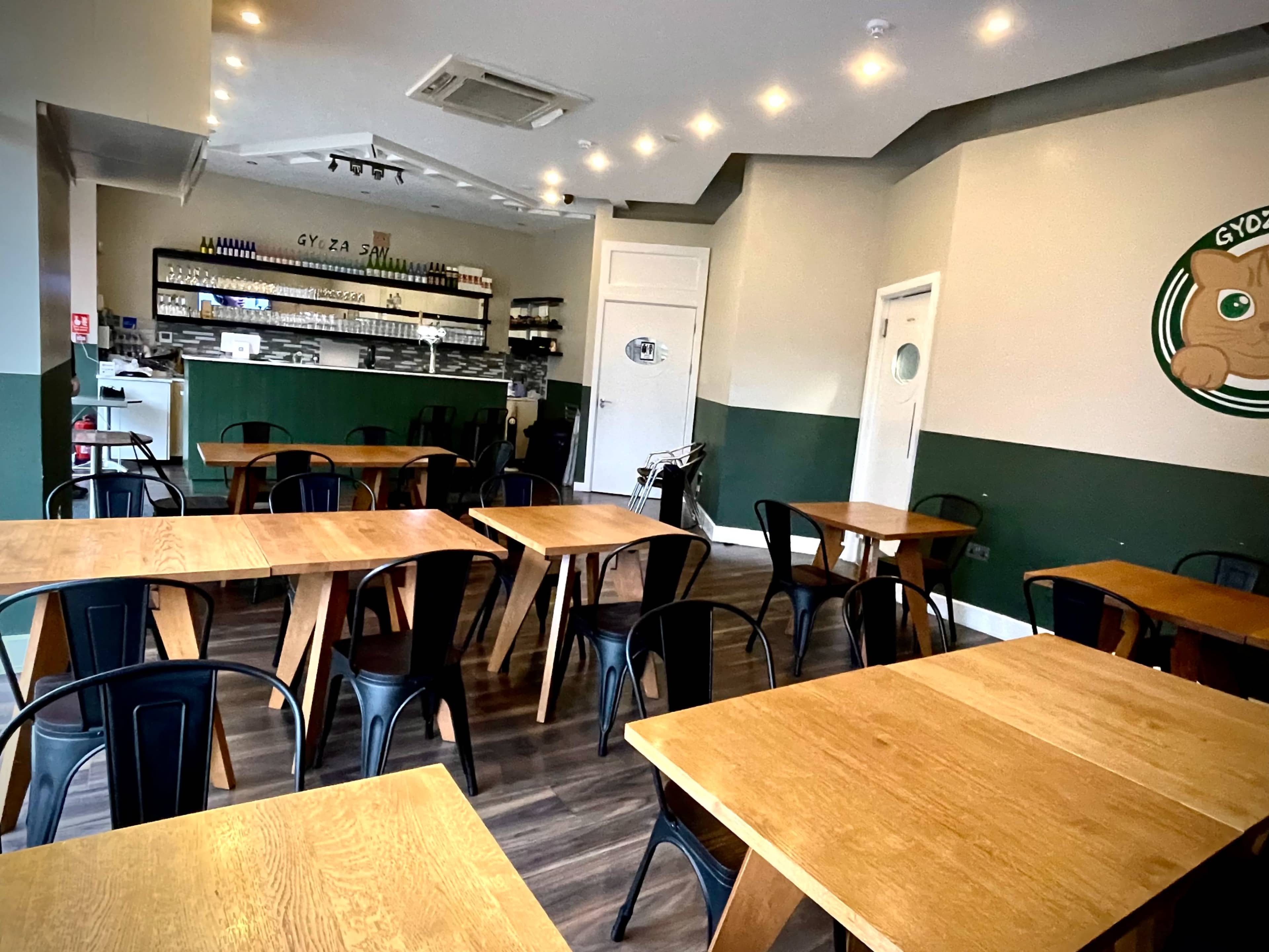 A neatly arranged restaurant interior featuring wooden tables and black metal chairs, with a service counter and shelves in the background.