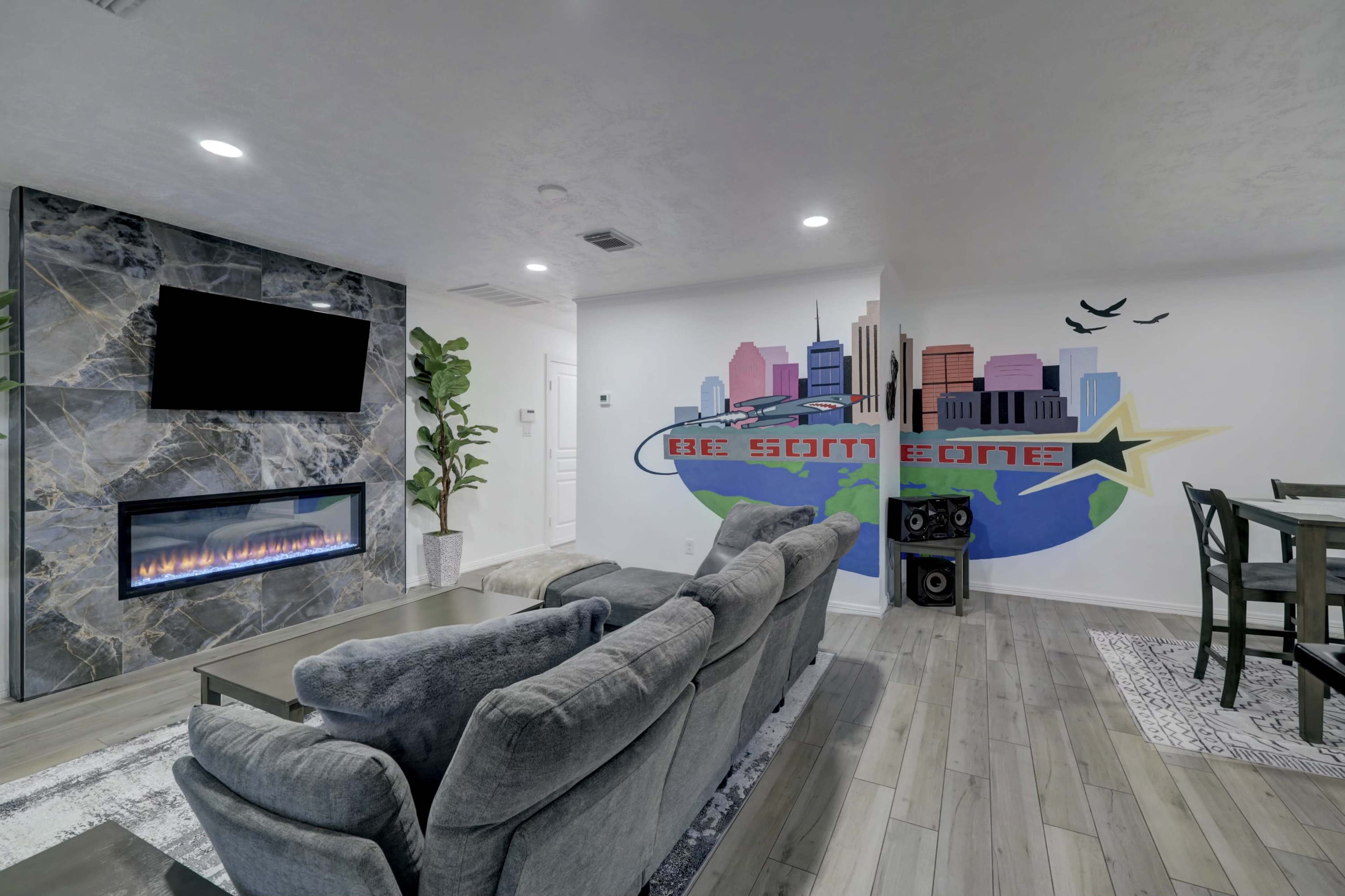 A modern living room features a sleek fireplace, a large TV mounted on the wall, and a mural depicting a city skyline with the phrase "BE SOMEONE" beside it.