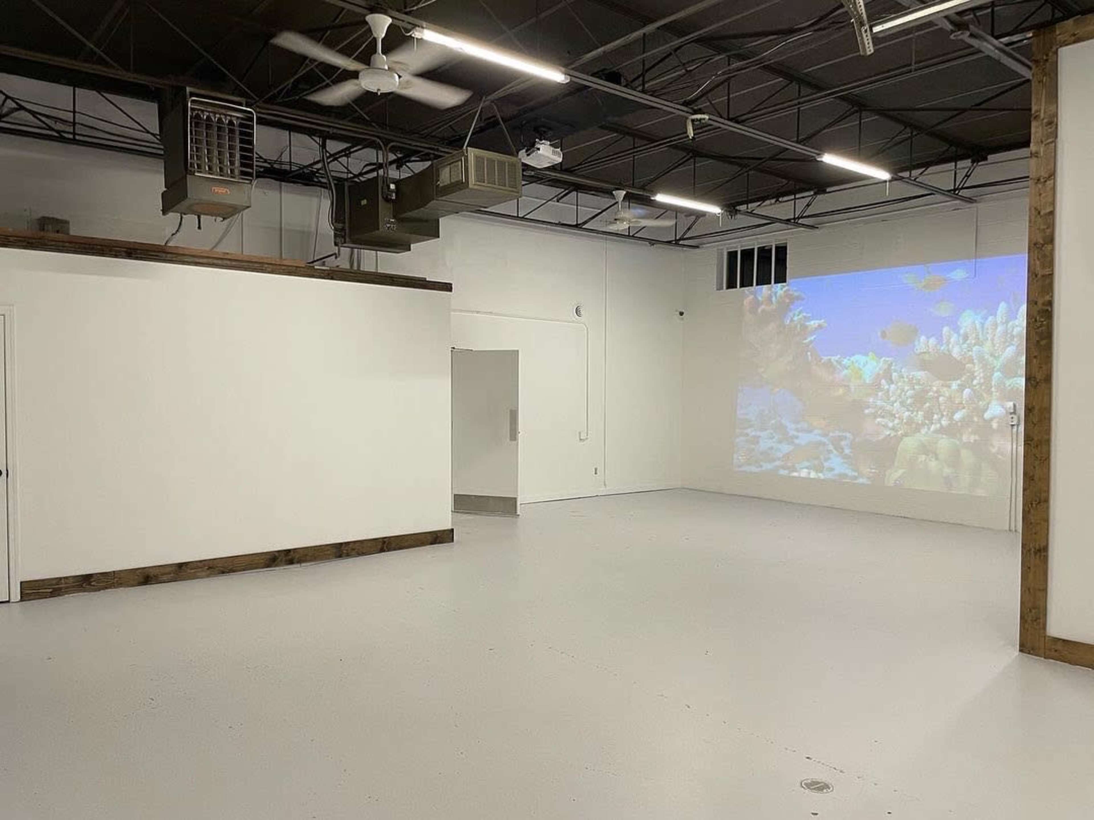 An empty room with a smooth white floor, a projection of a coral reef scene on one wall, and ventilation units mounted on the ceiling.