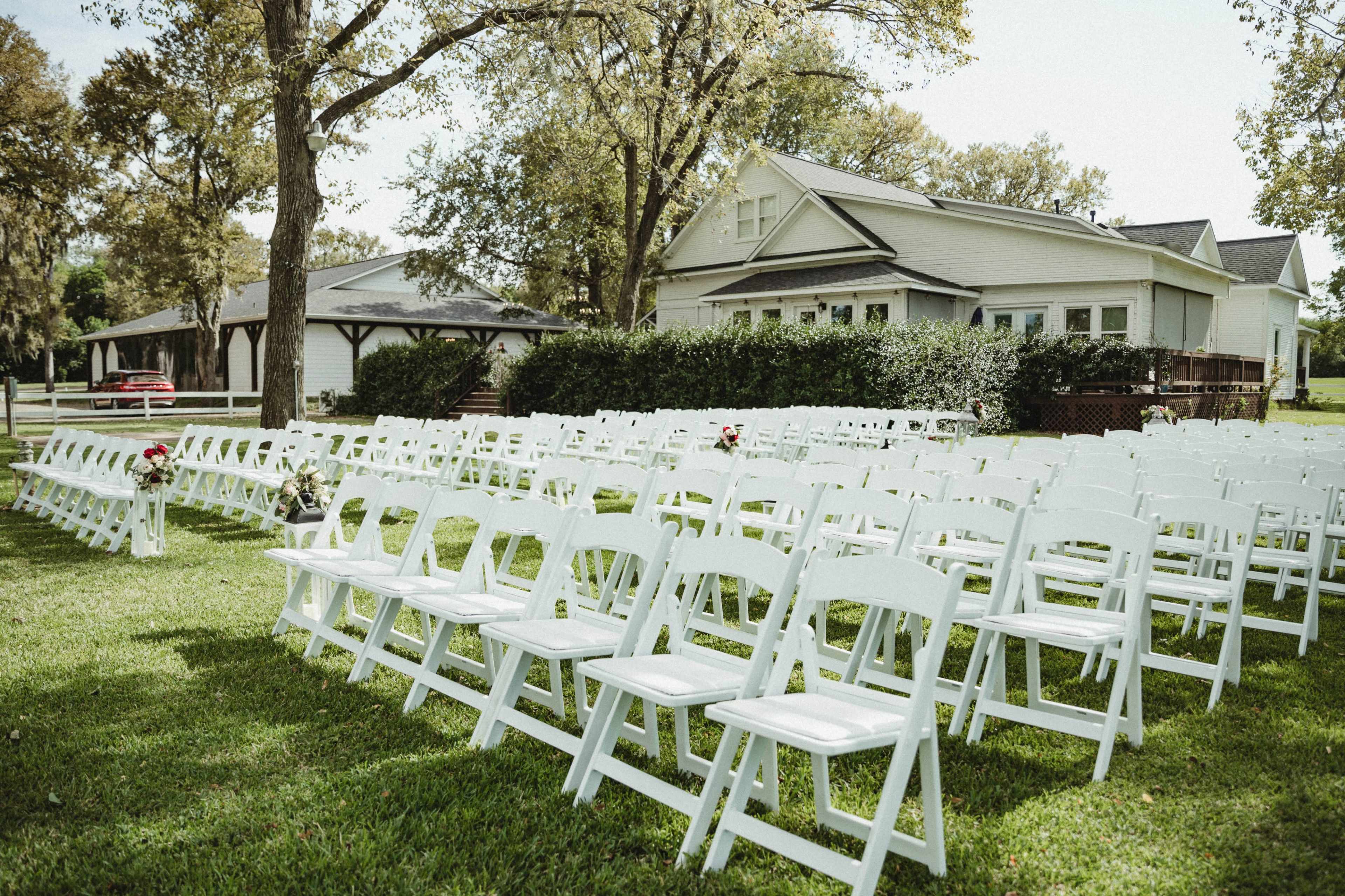 A row of white folding chairs is set up on a grassy area near a house, with trees and a fence in the background.