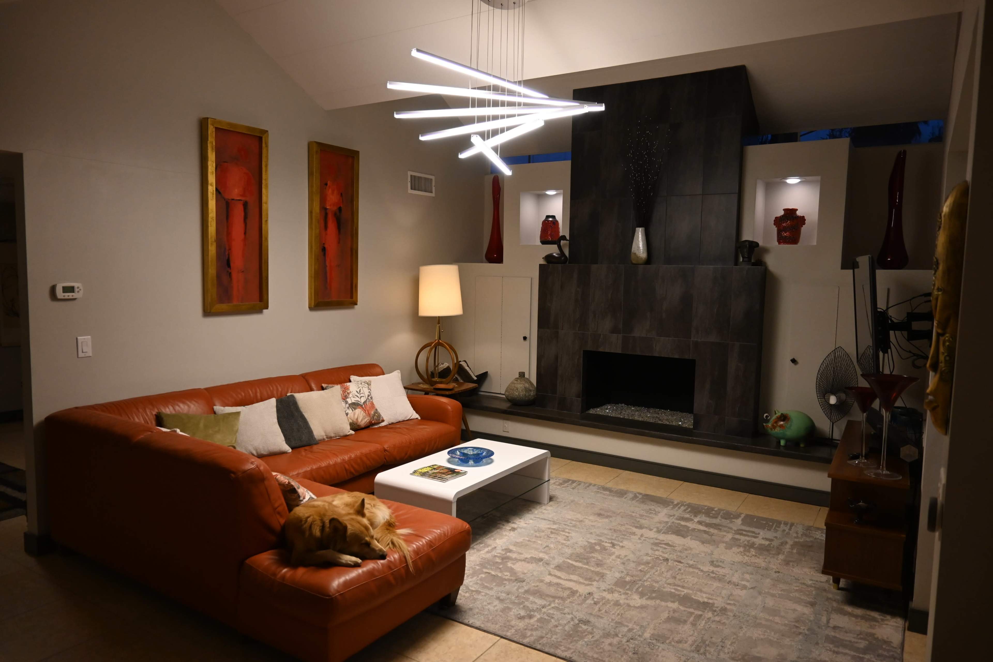 A modern living room features an orange sectional sofa, a white coffee table, and a contemporary light fixture hanging above a stone fireplace, with decorative art on the walls.