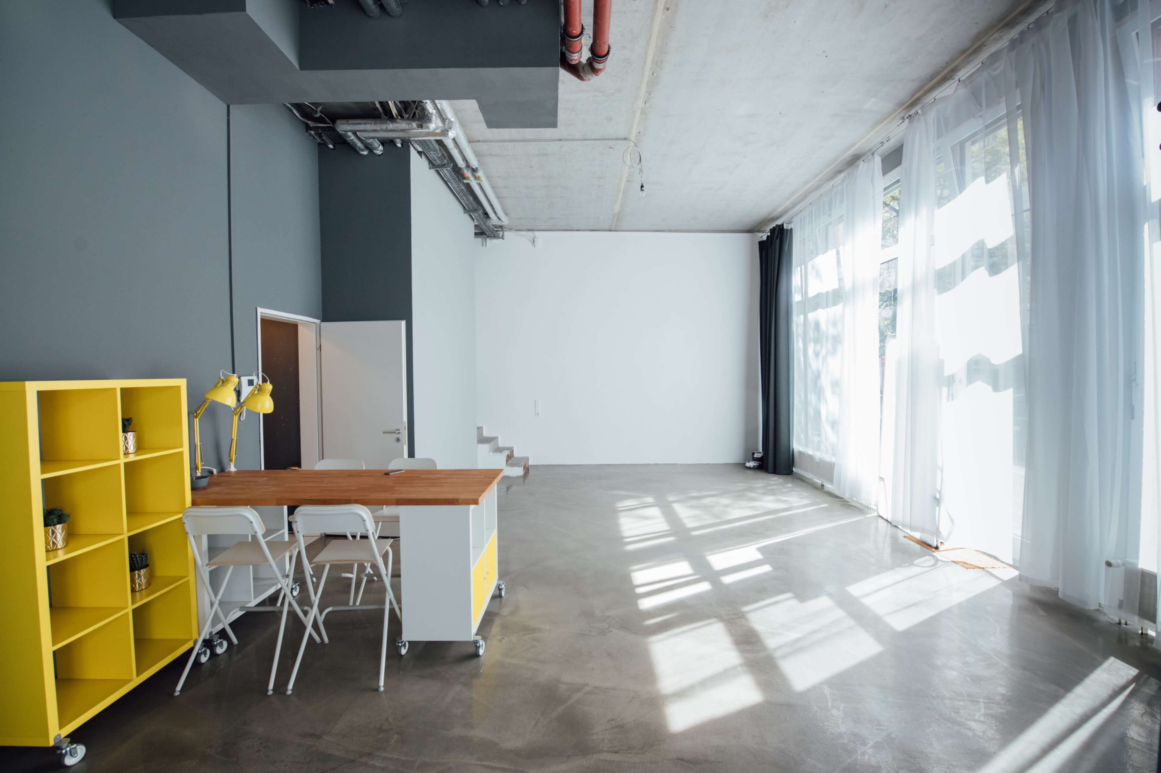 The image features a spacious, modern room with large windows allowing natural light to fill the area, a concrete floor, and a yellow shelving unit with a small countertop and stools.