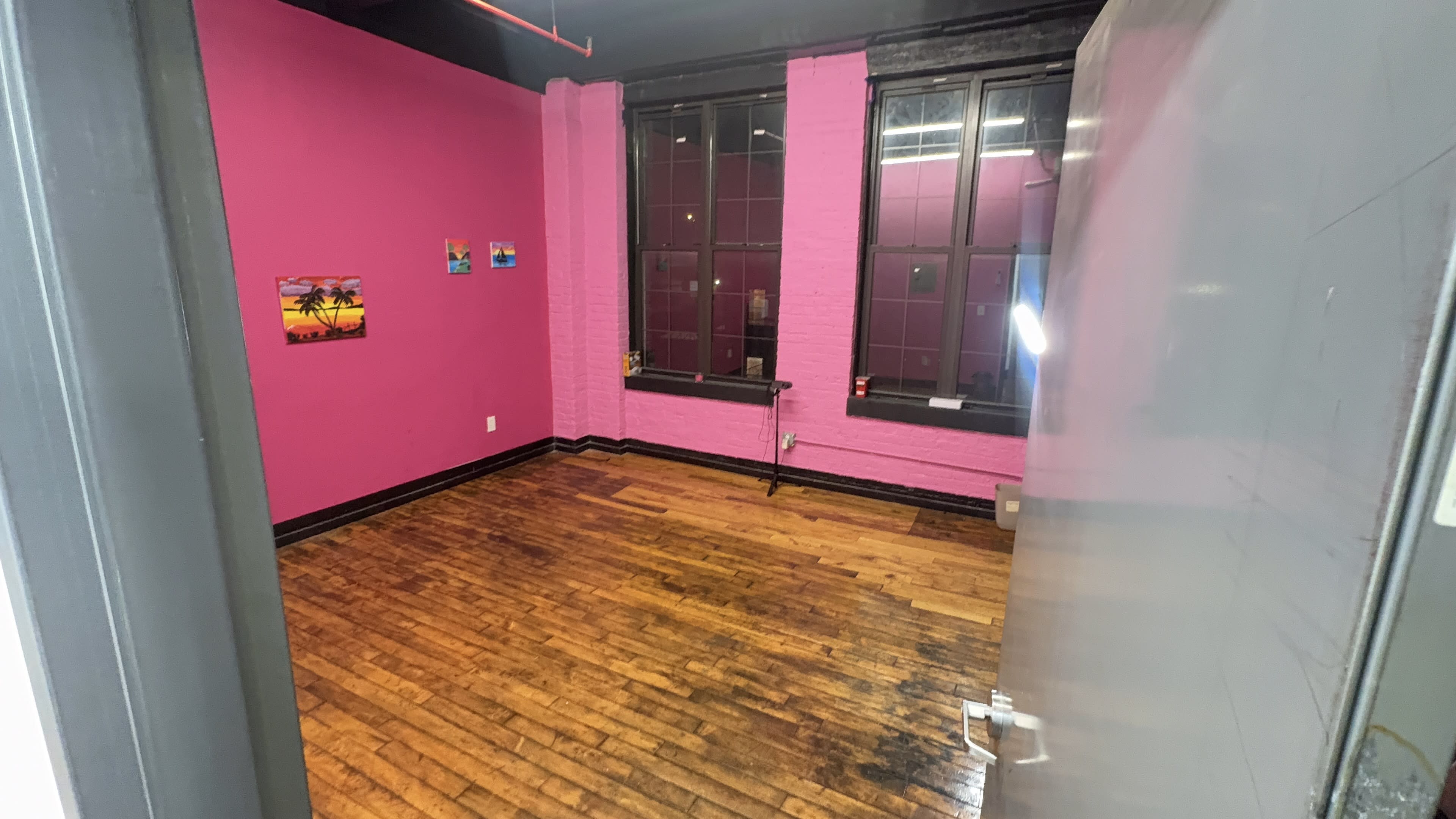 The image shows an empty room with pink walls, large windows, and wooden flooring.