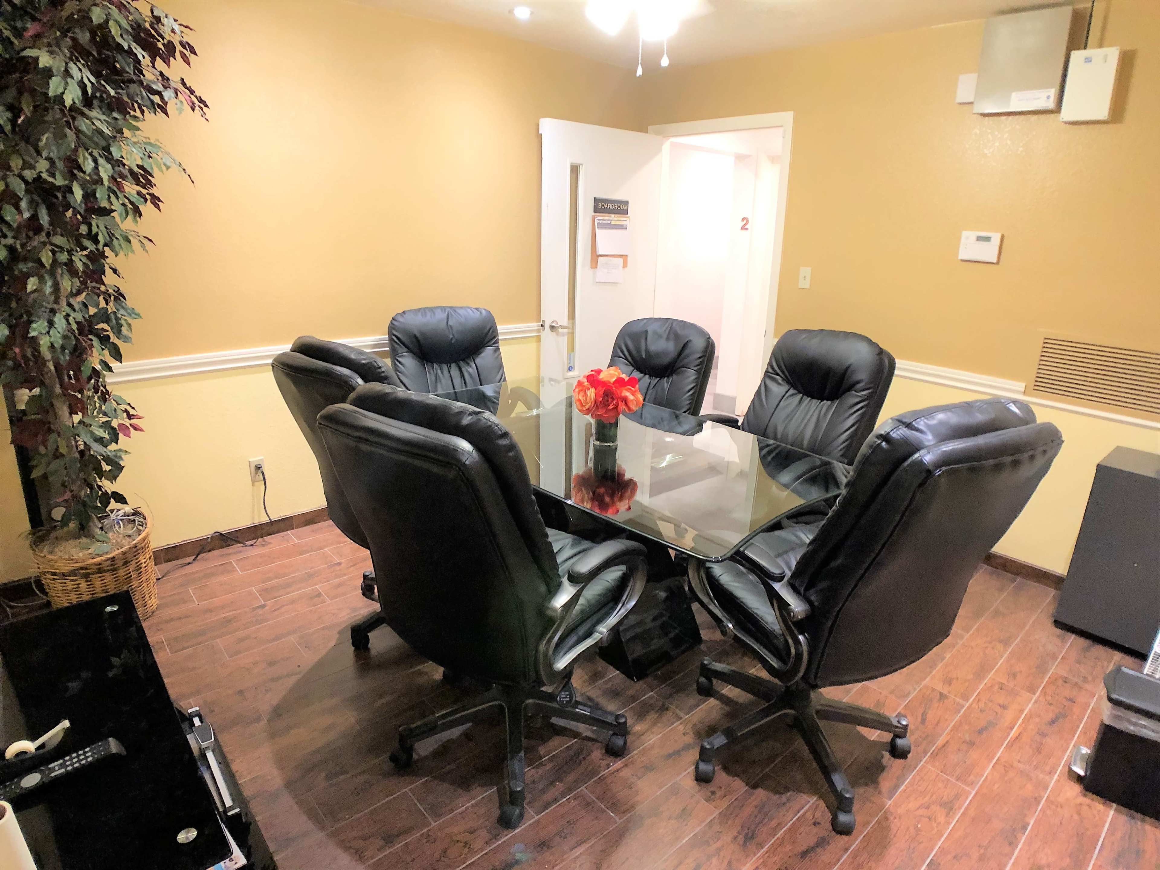 The image shows a conference room with a glass-topped circular table surrounded by six black leather office chairs and a decorative plant in the corner.