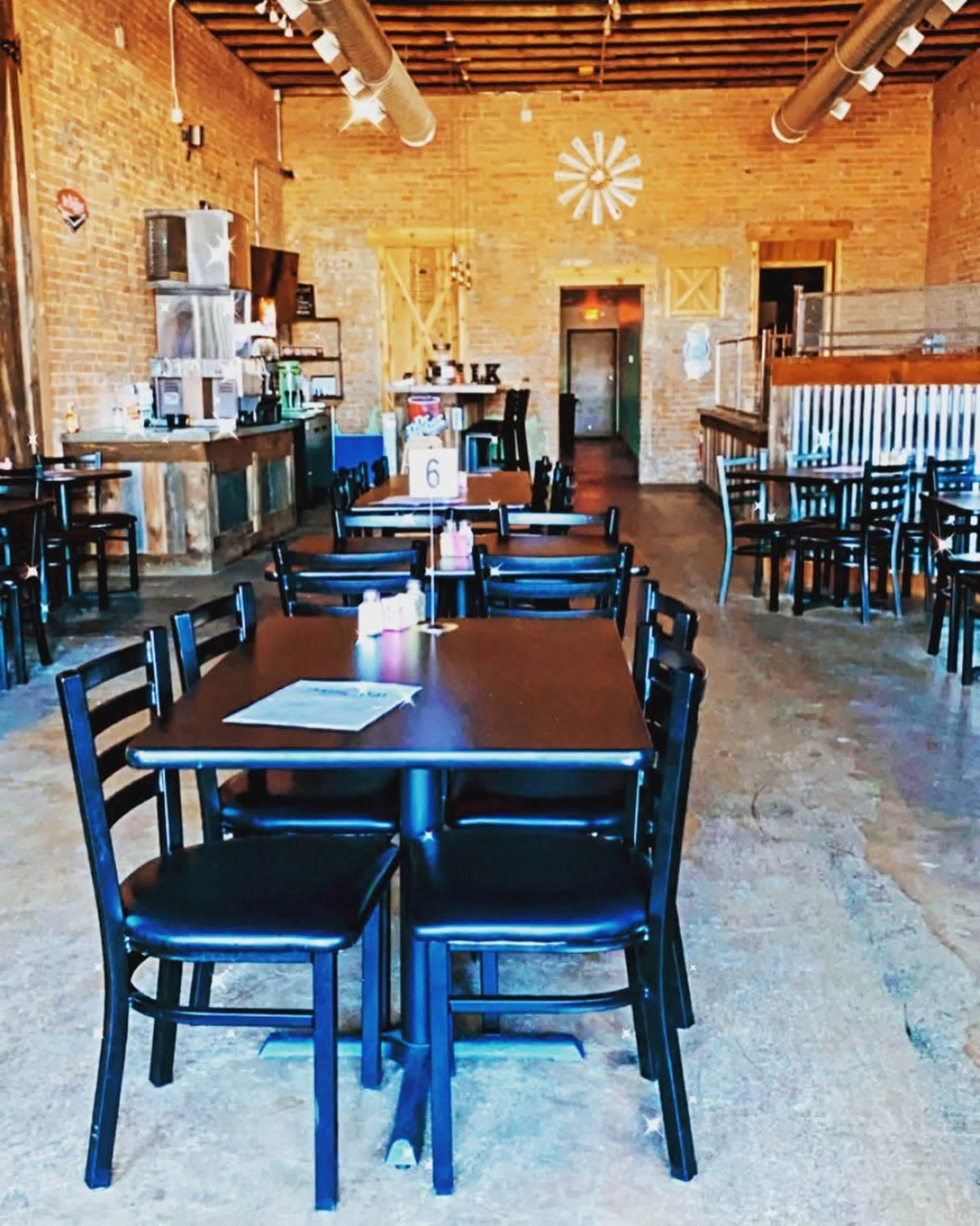 The image shows an empty restaurant interior with wooden floors, tables set for dining, and a rustic bar area in the background.