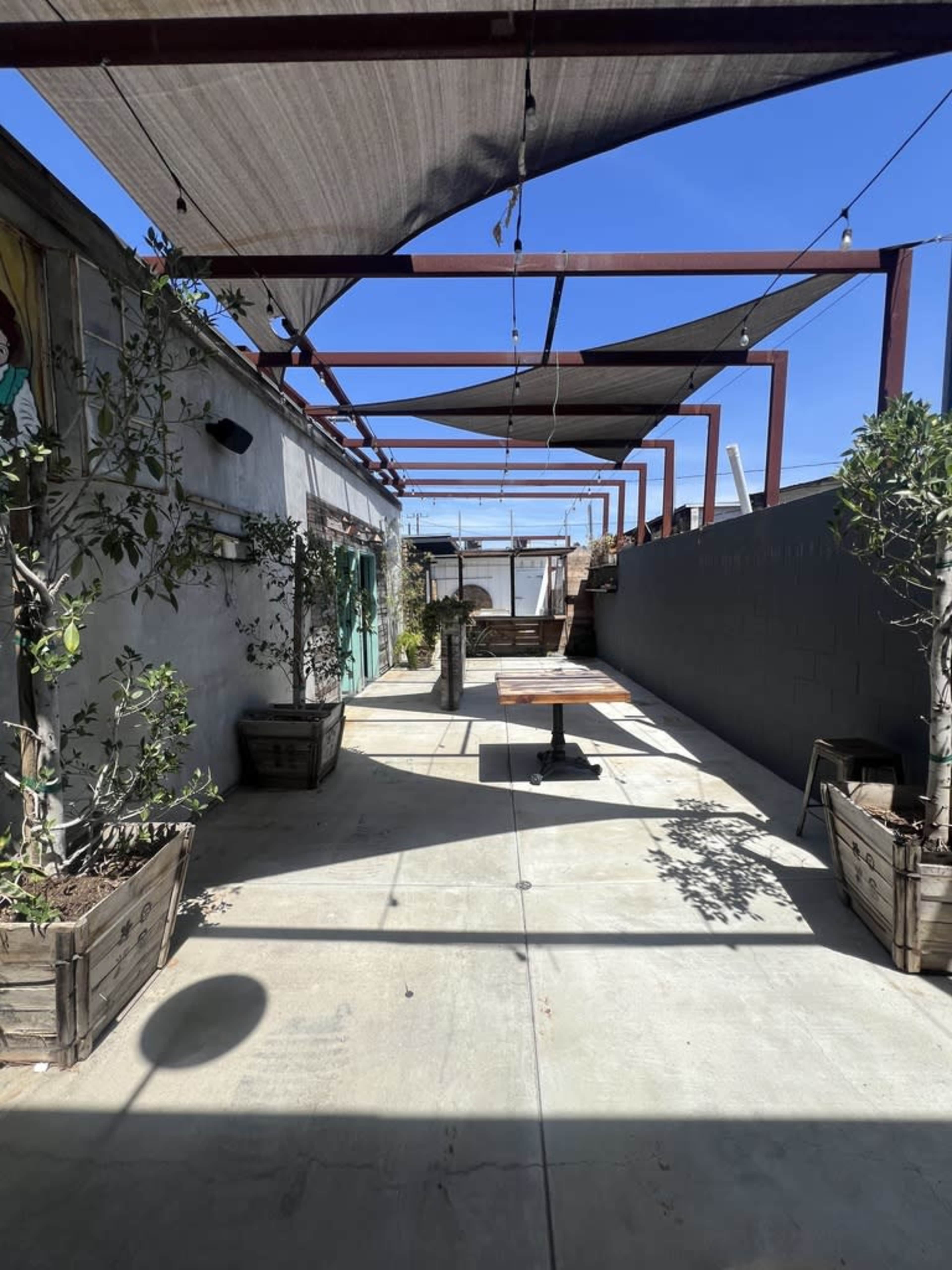 The image shows a long, narrow outdoor space with shaded pergolas, potted plants, and a concrete floor, leading towards a distant open area.