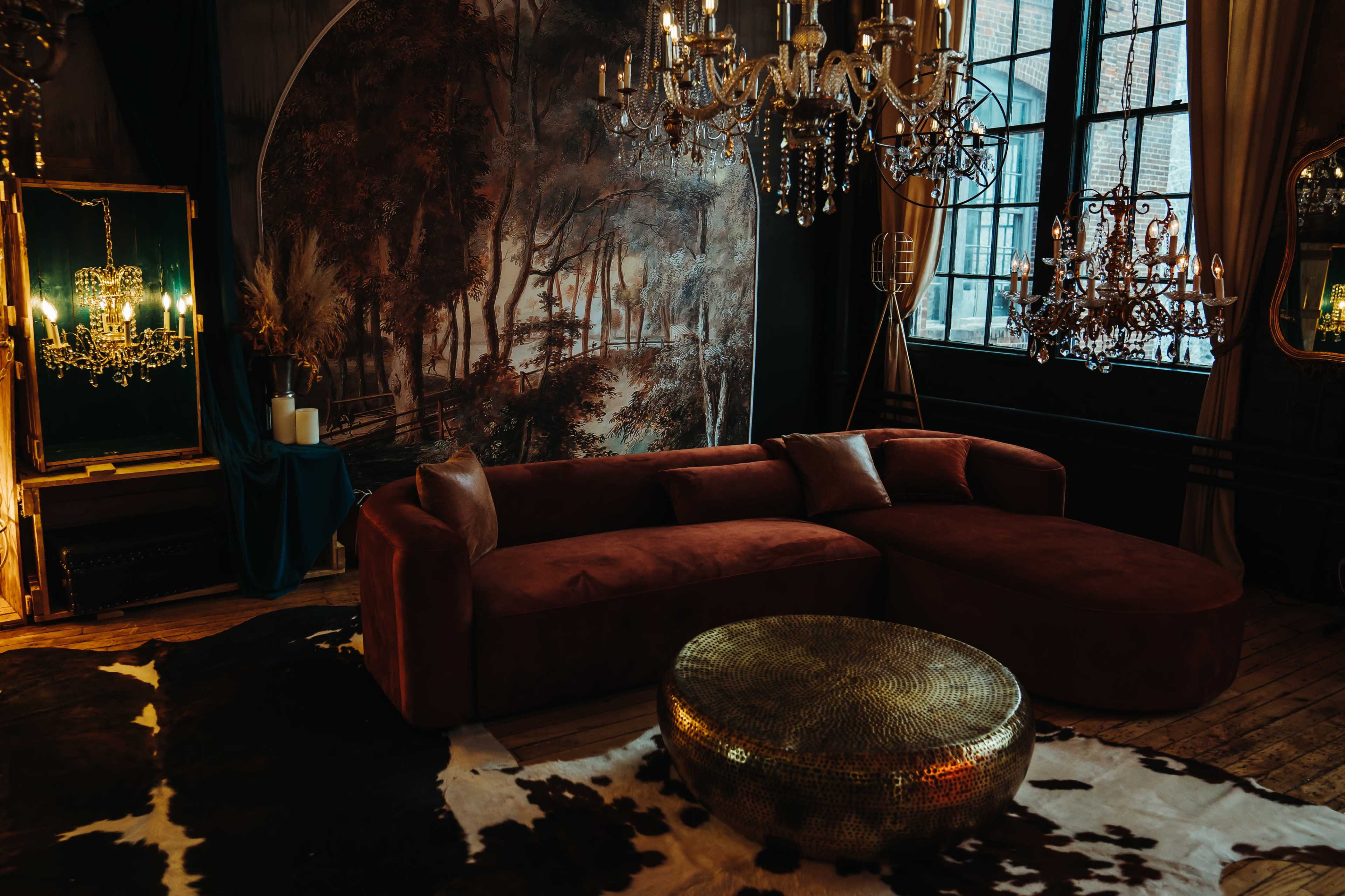 The scene features a stylish lounge area with a curved maroon sofa, a round gold coffee table, and elegant chandeliers, all set against a backdrop of a scenic mural.