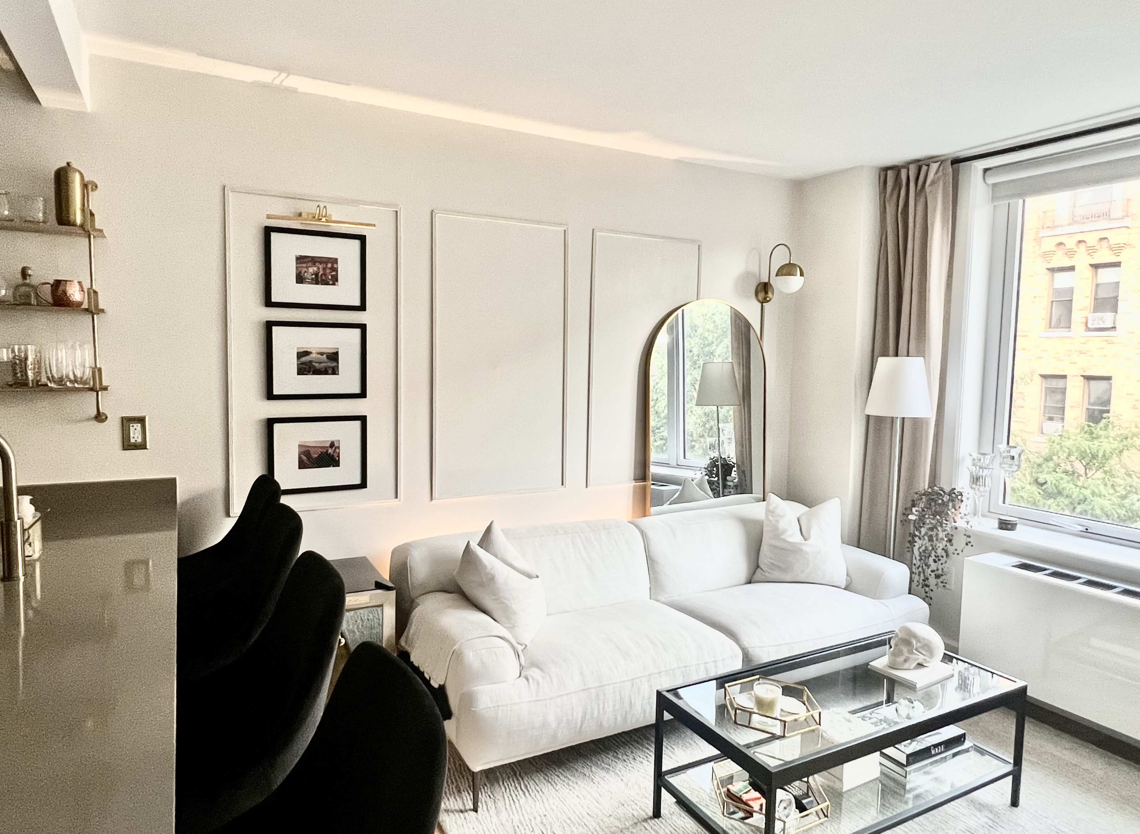 A modern living room features a white sofa, a glass coffee table, framed pictures on the wall, and large windows with a view of greenery outside.