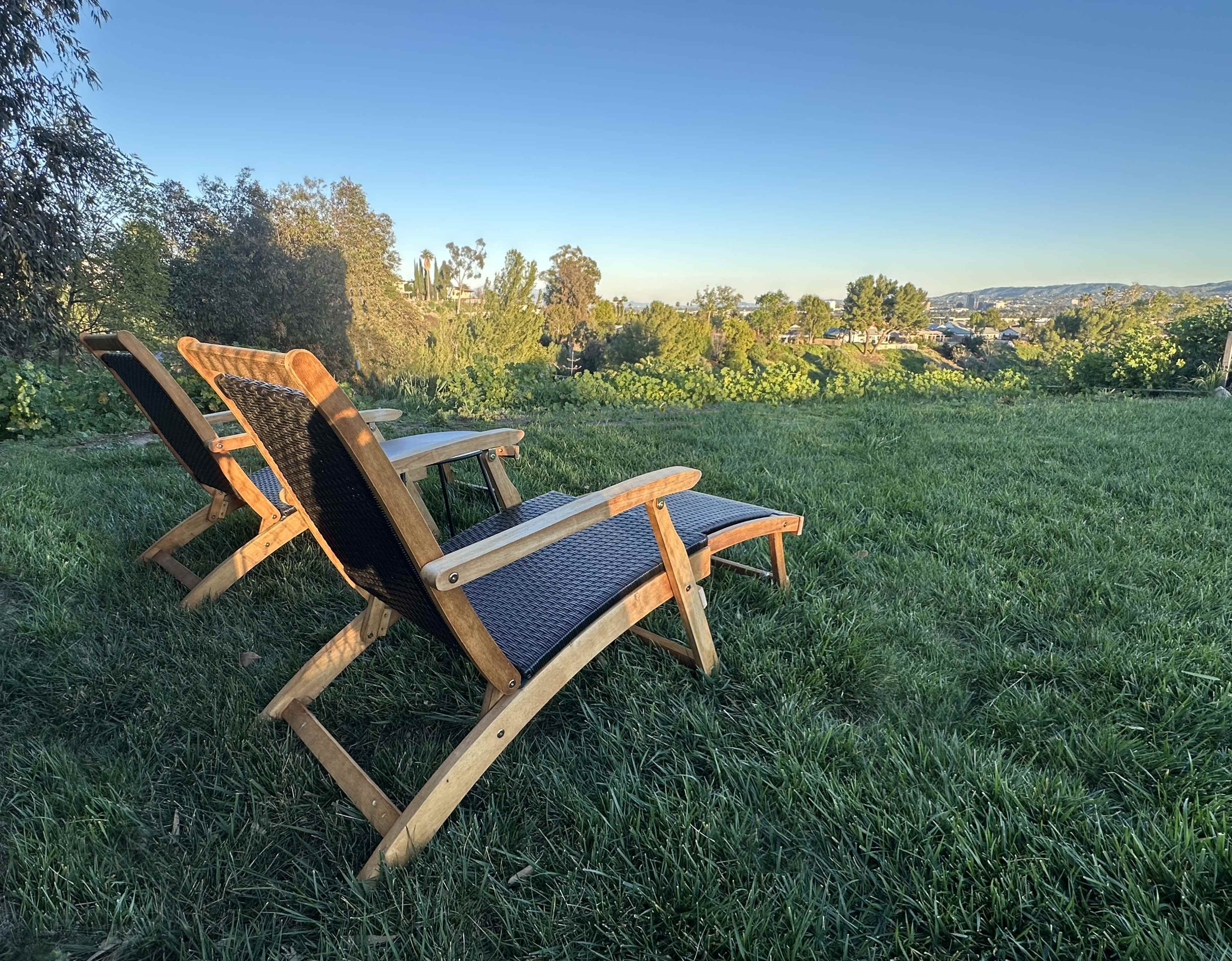 Three wooden lounge chairs are arranged on a grassy area, facing a landscape of greenery and distant hills under a clear blue sky.