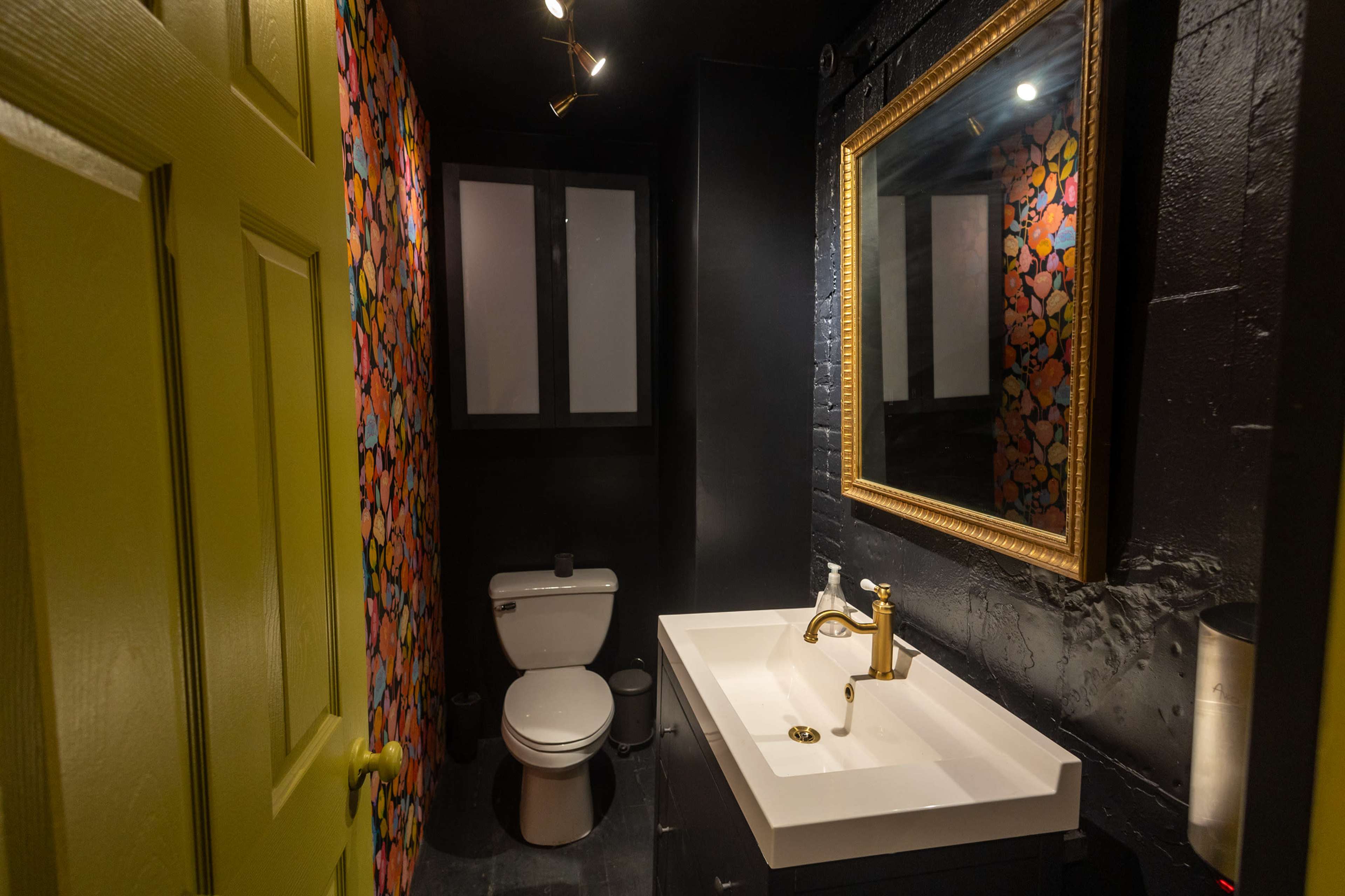 A compact bathroom featuring a toilet, sink with a gold faucet, and colorful wallpaper, illuminated by track lighting.
