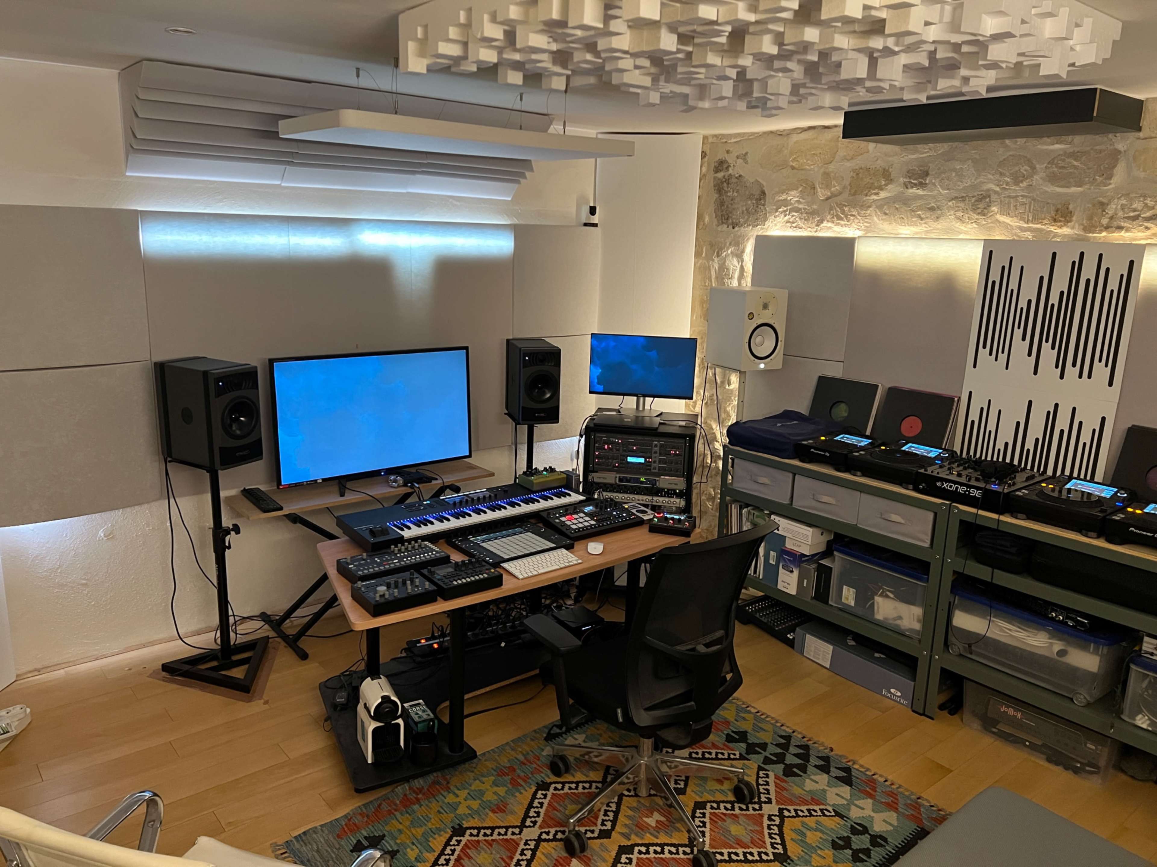 The image shows a modern music production studio with a computer setup, MIDI keyboard, audio speakers, and various electronic instruments organized on shelves.