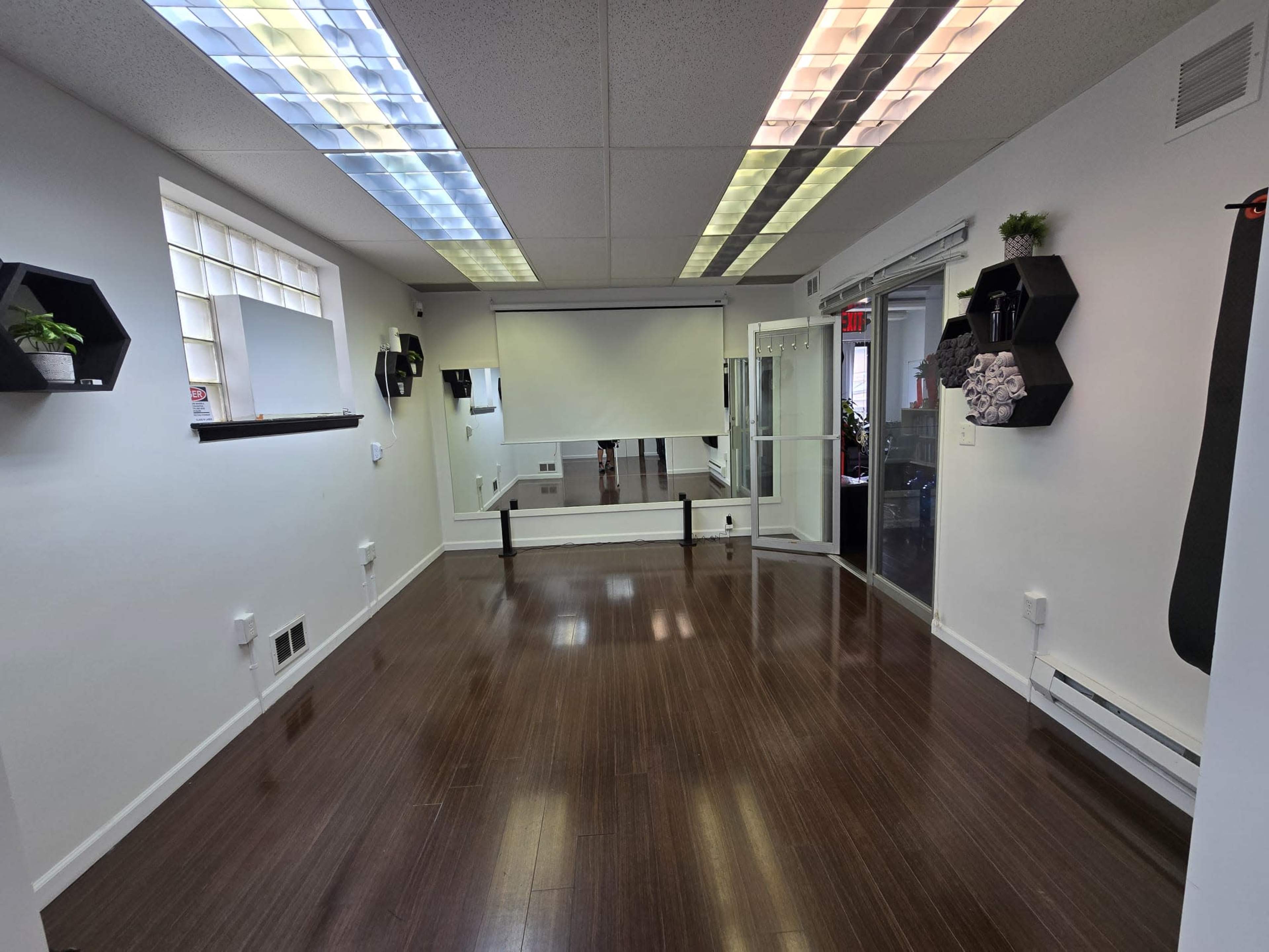 A spacious, minimalist room with wooden flooring, a projector screen on the far wall, and decorative black hexagonal shelves.