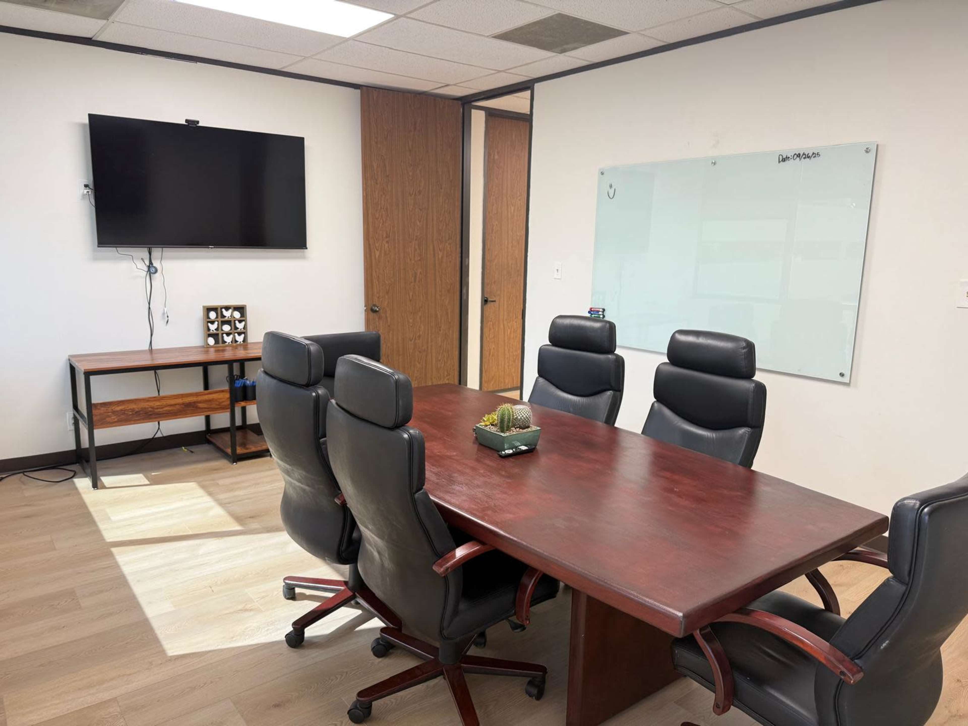 The image shows a conference room with a large wooden table surrounded by six black leather chairs, a wall-mounted TV, and a whiteboard.