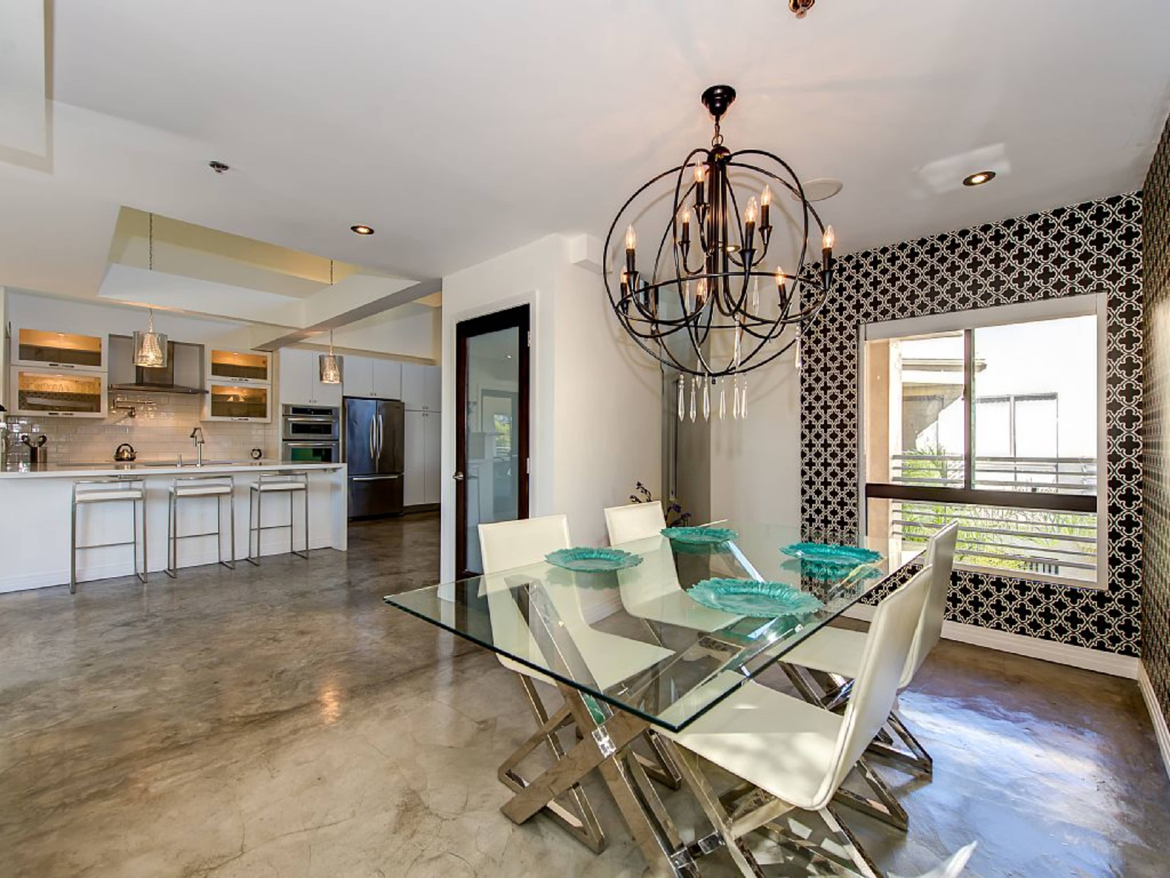 A modern dining area features a glass table with white chairs and a chandelier, adjacent to a kitchen with stainless steel appliances.