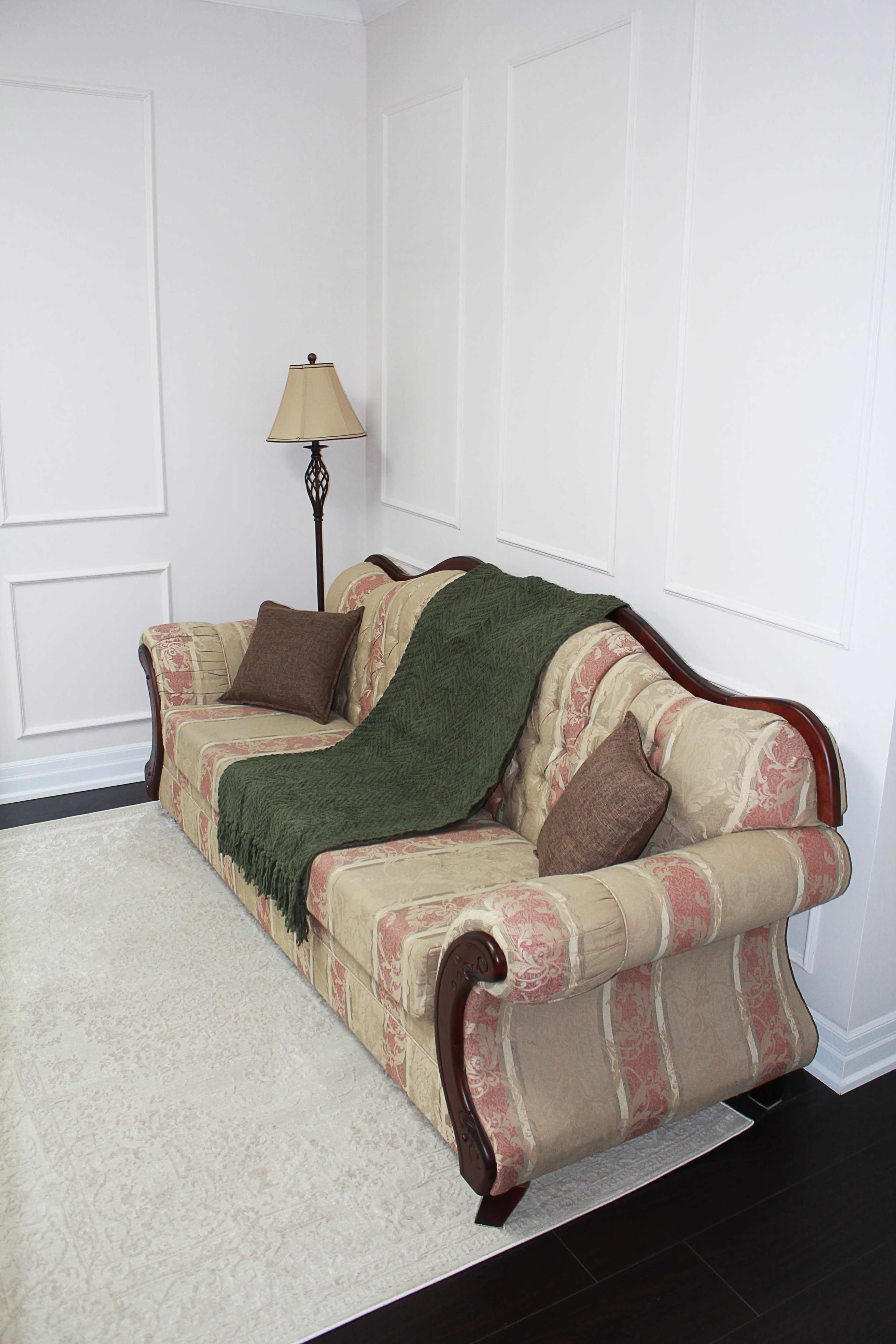 A patterned sofa with decorative cushions and a green throw blanket is positioned beside a floor lamp against a light-colored wall.