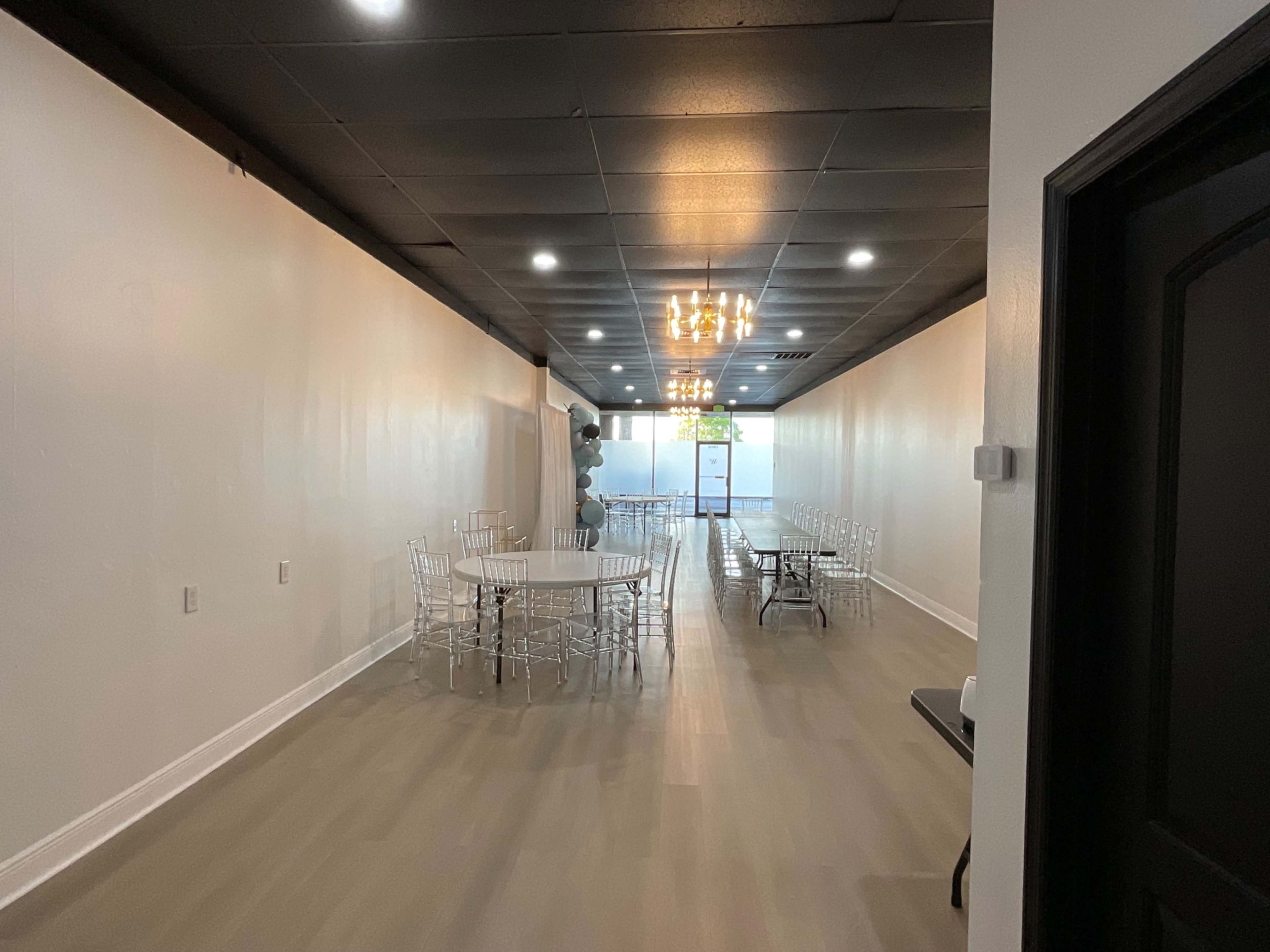 A spacious, well-lit room with tables and chairs arranged in a seating area, featuring a chandelier on the ceiling.