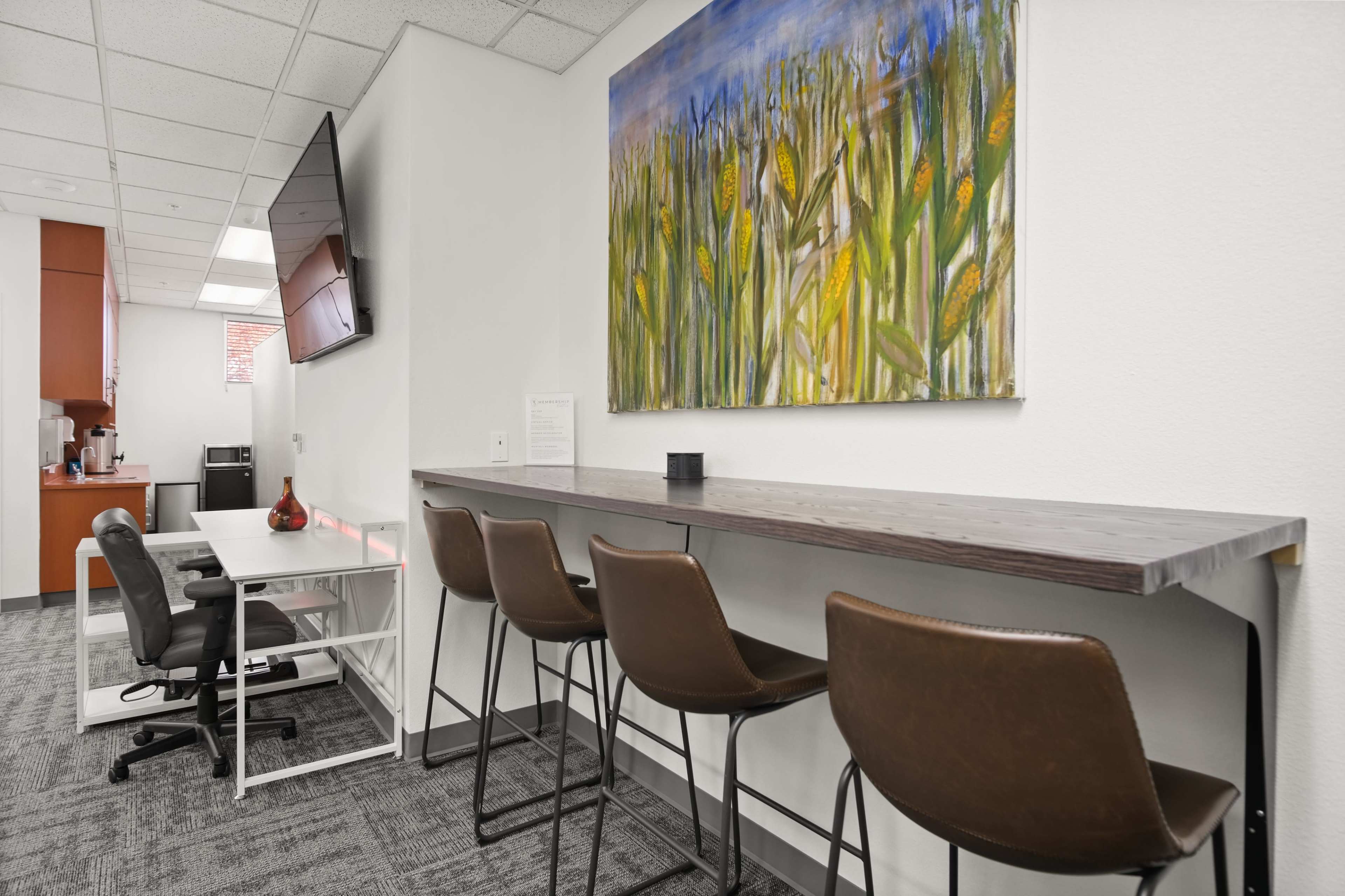 A modern workspace featuring a bar-style table with four brown stools, a black chair at a desk, and a colorful painting of cornfields on the wall.