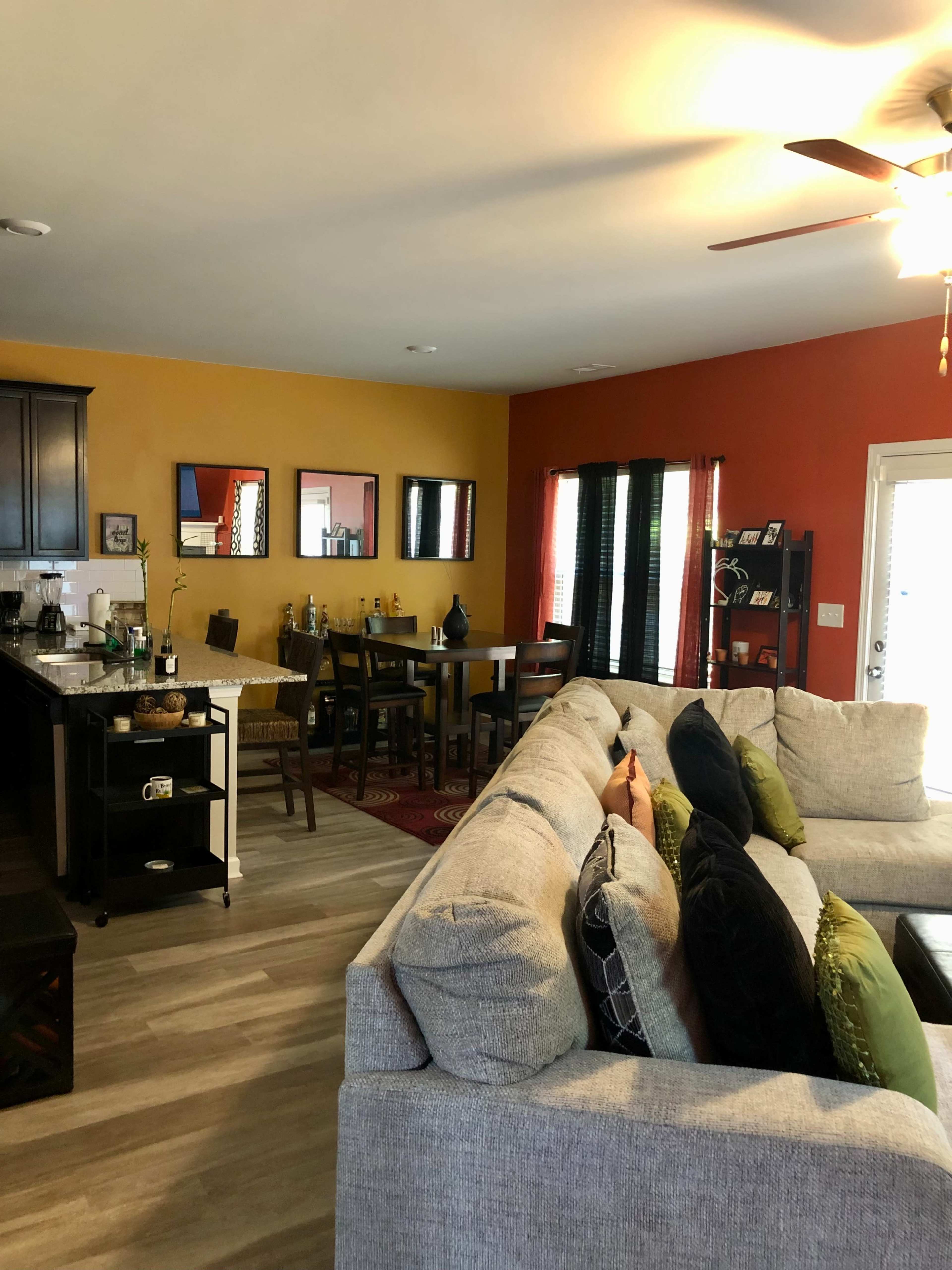 A spacious living area features a sectional sofa facing a kitchen with dark cabinets, a dining table, and colorful accent walls.