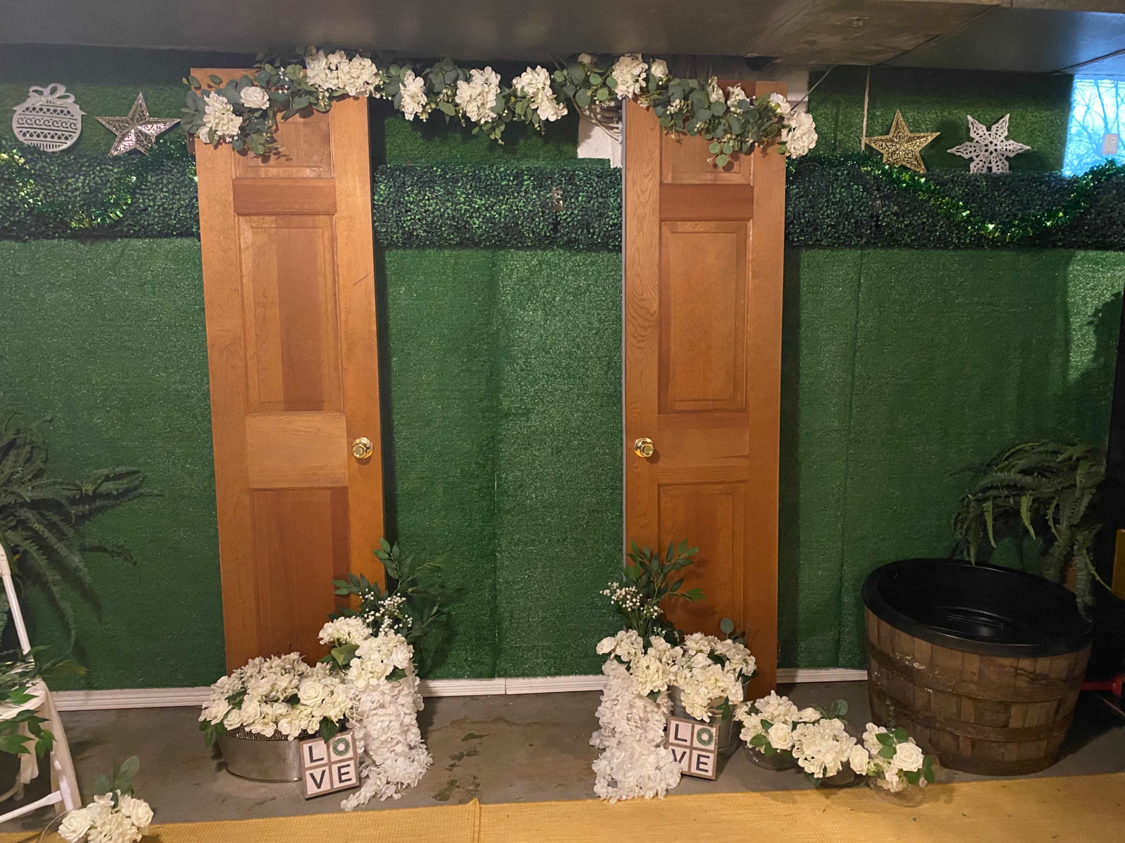 The image shows two wooden doors adorned with greenery and white flowers, flanked by decorative elements like stars and a barrel in a basement setting.