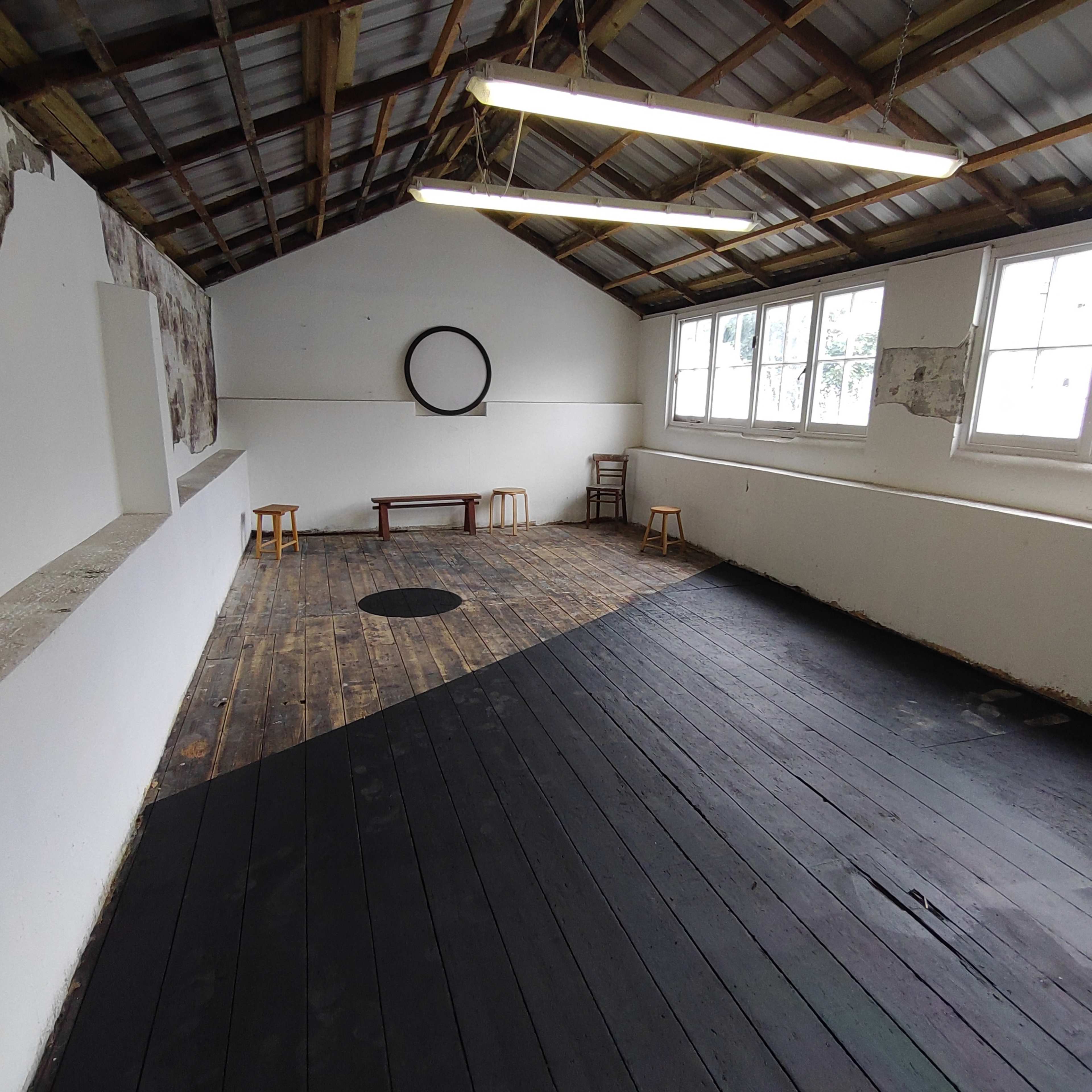 An empty room with a wooden floor, white walls, a circular mirror on the wall, and several stools arranged throughout the space.