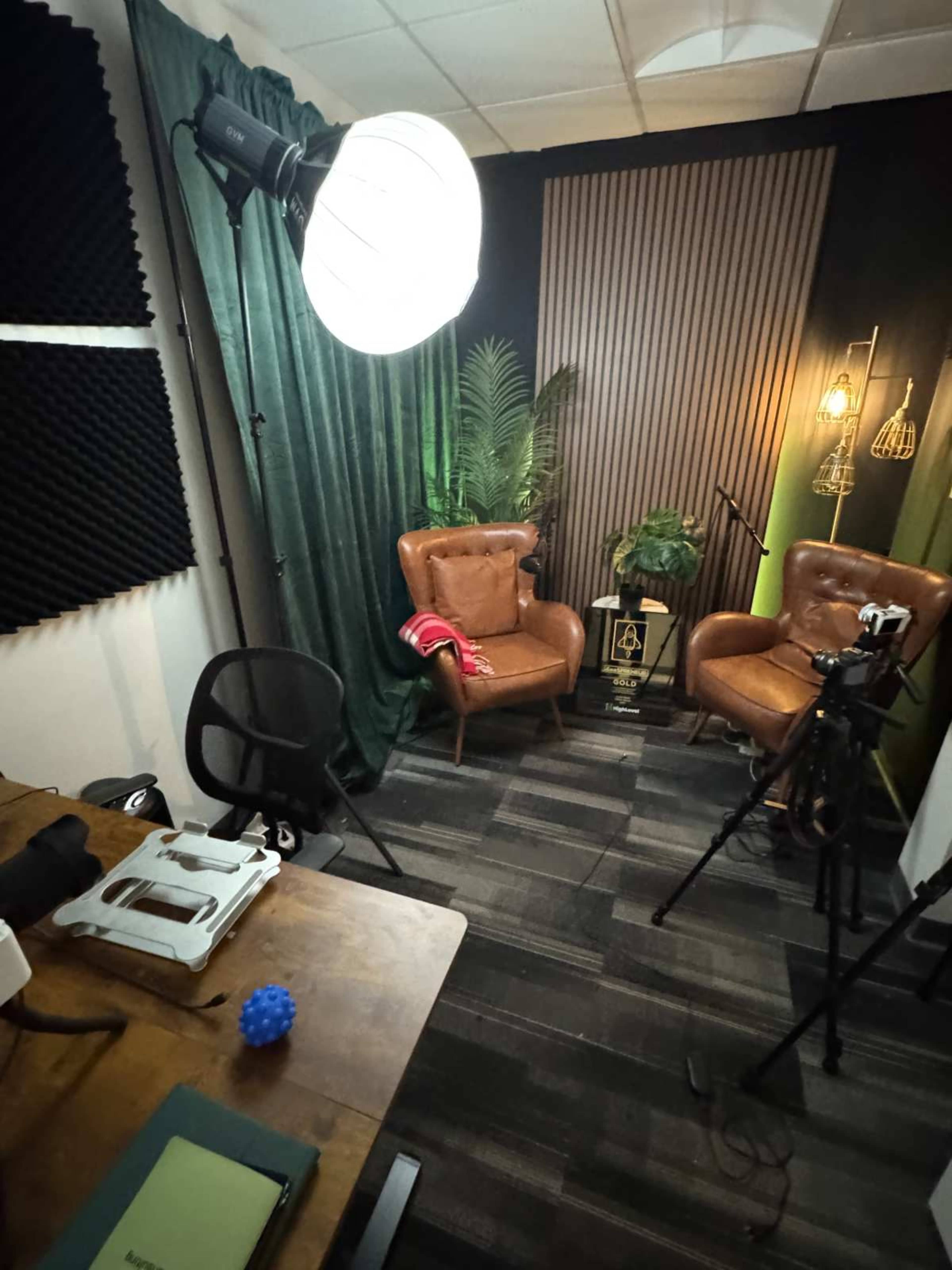 The image shows a small recording studio setup featuring two brown chairs, a green backdrop, and various lighting and filming equipment.