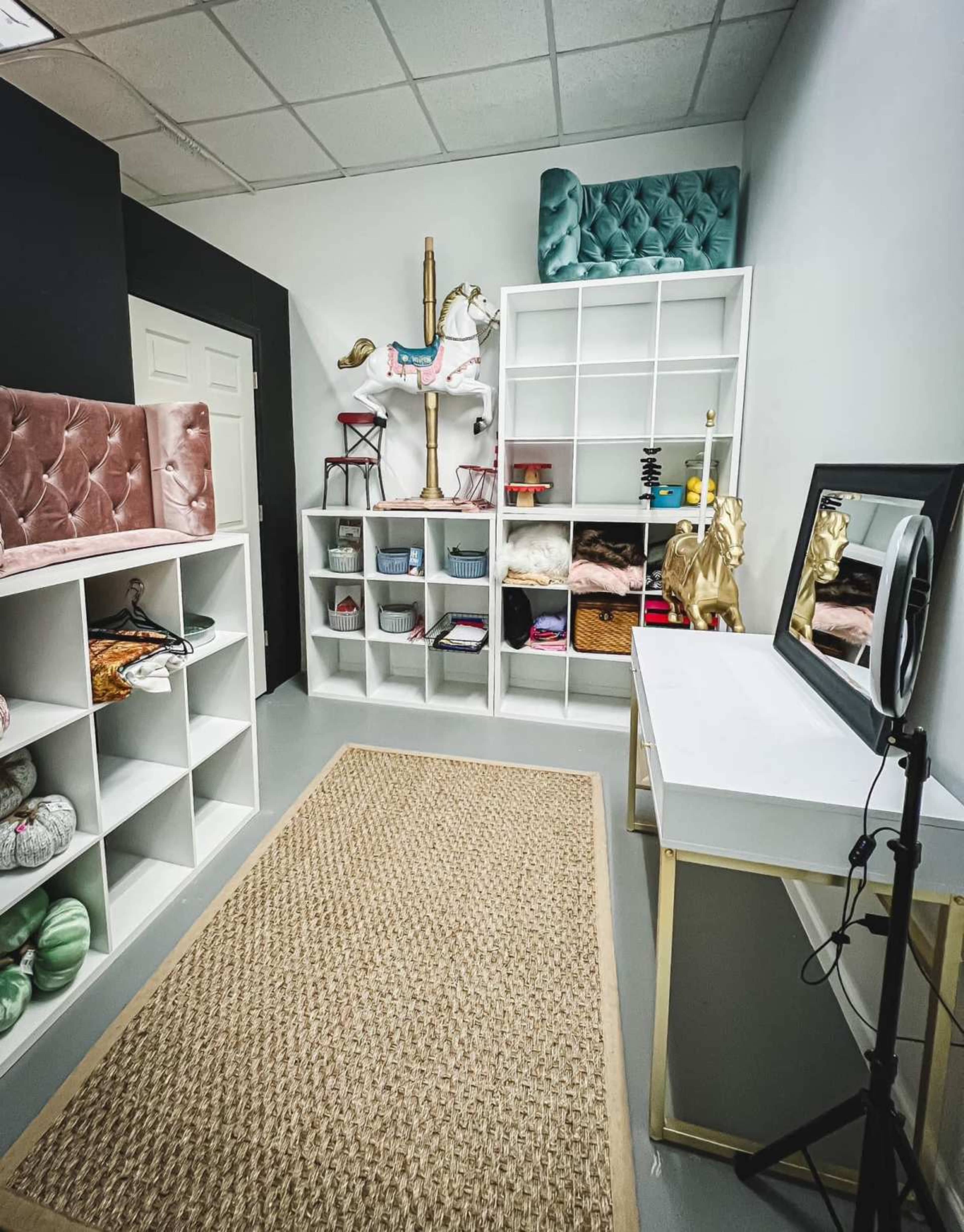 The image shows a neatly organized room with white shelving units filled with various colored fabrics and baskets, a mirrored vanity with a ring light, and decorative elements including a gold statue and a plush chair.