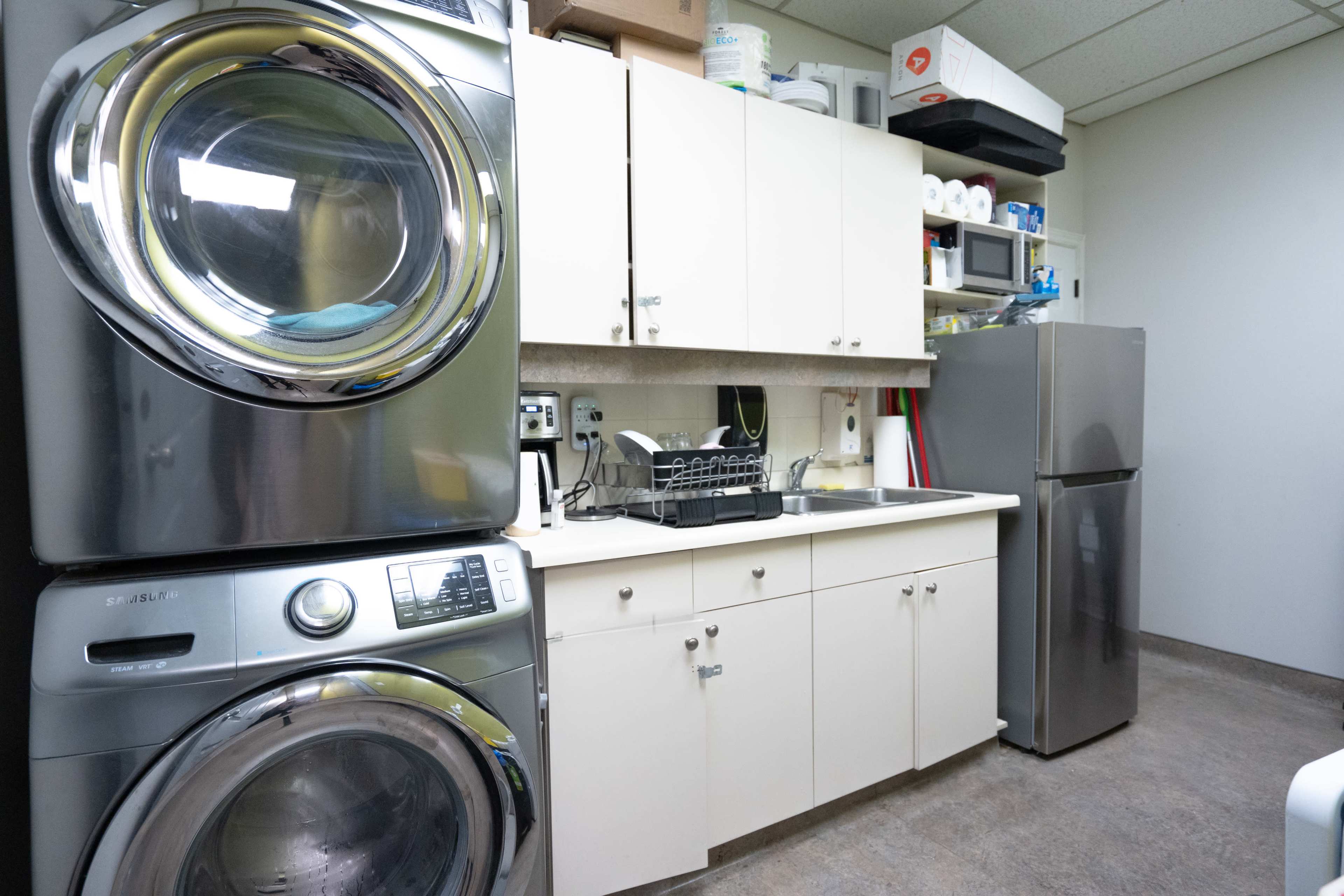 The image shows a laundry room featuring stacked washing and drying machines, a countertop with a dish rack, and cabinets above.