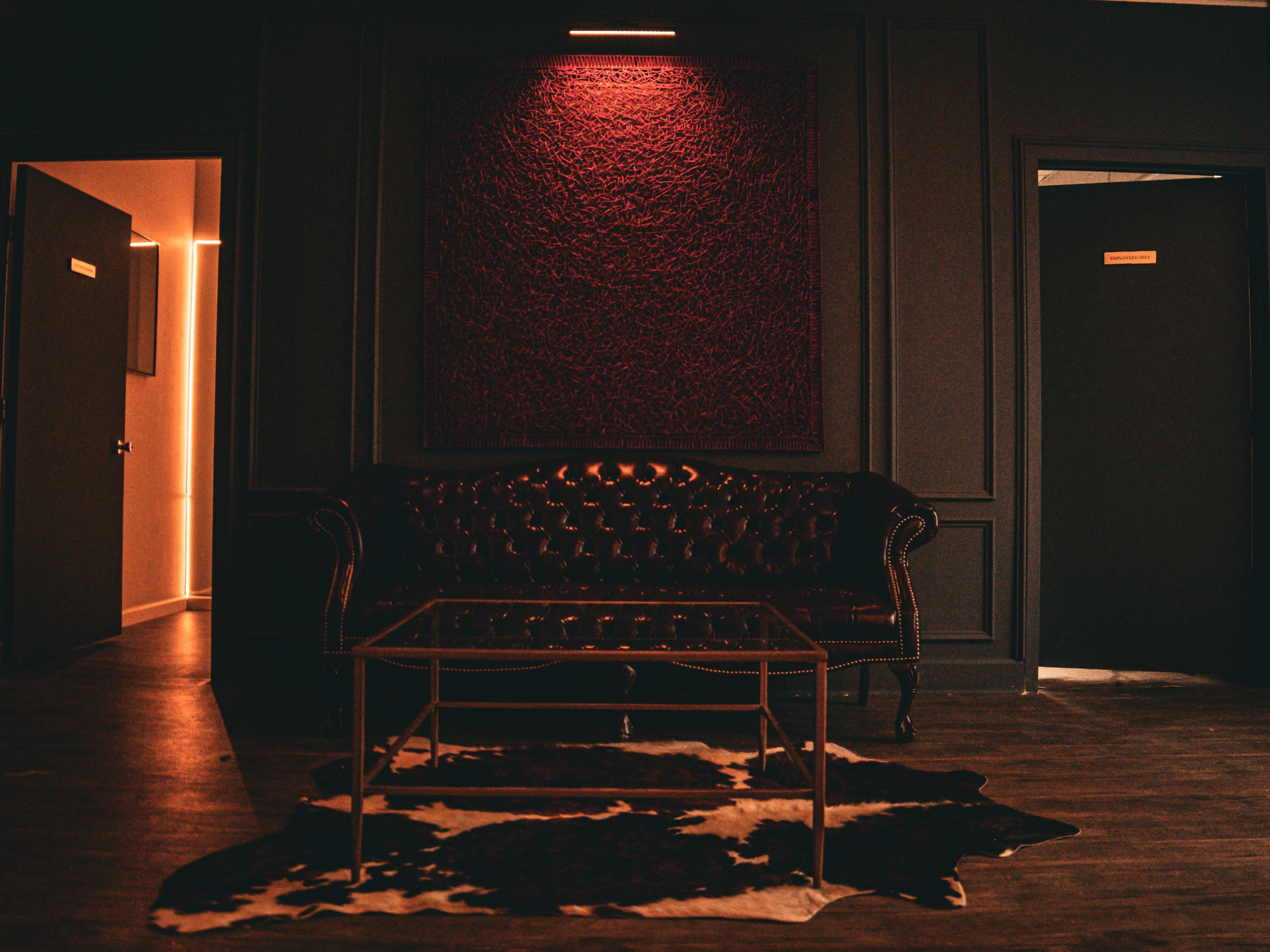 The image shows a dimly lit room featuring a dark leather sofa, a glass coffee table, and a cowhide rug on a wooden floor.