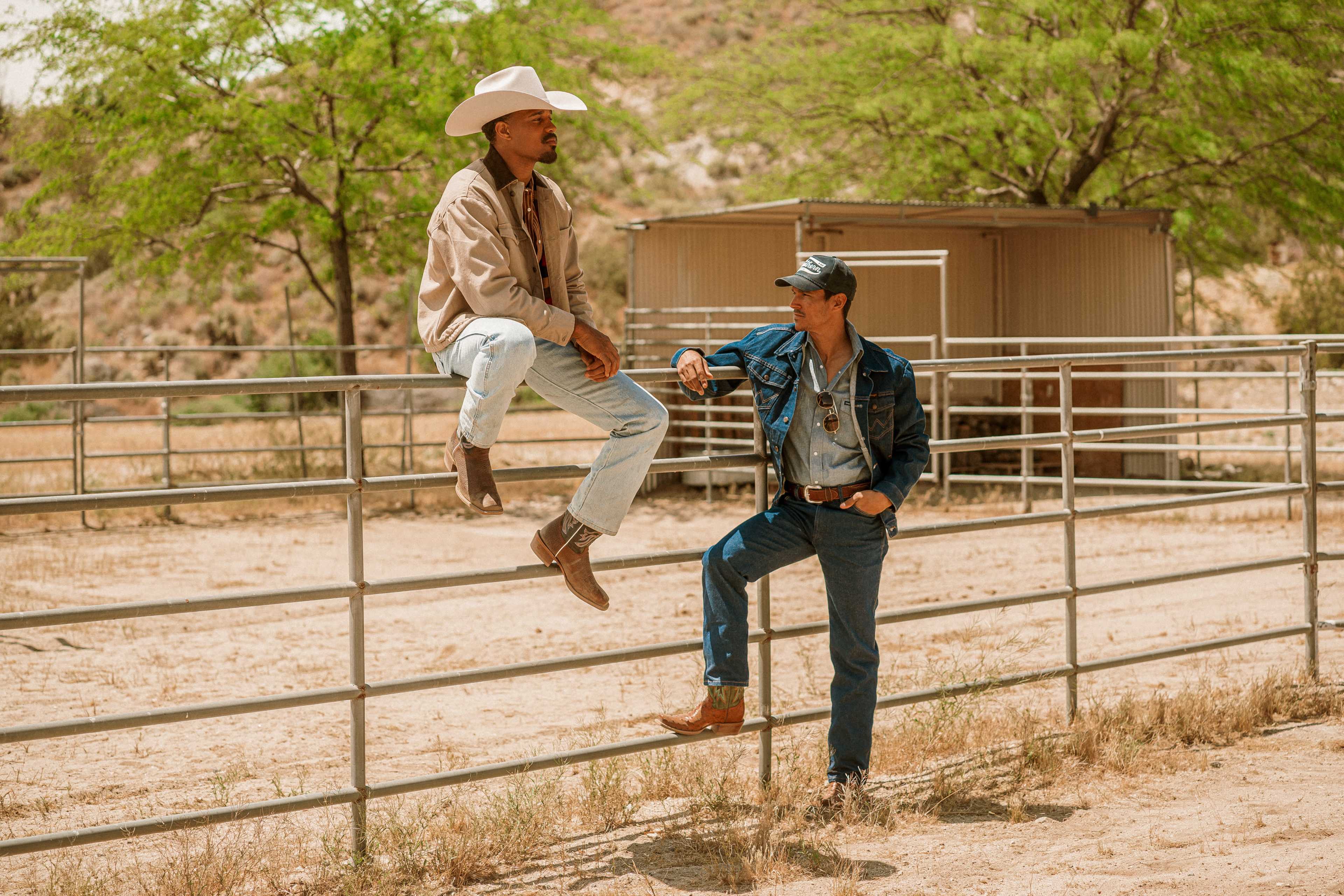 Two men, one wearing a cowboy hat and the other in a denim jacket, are sitting on a fence in a ranch setting.