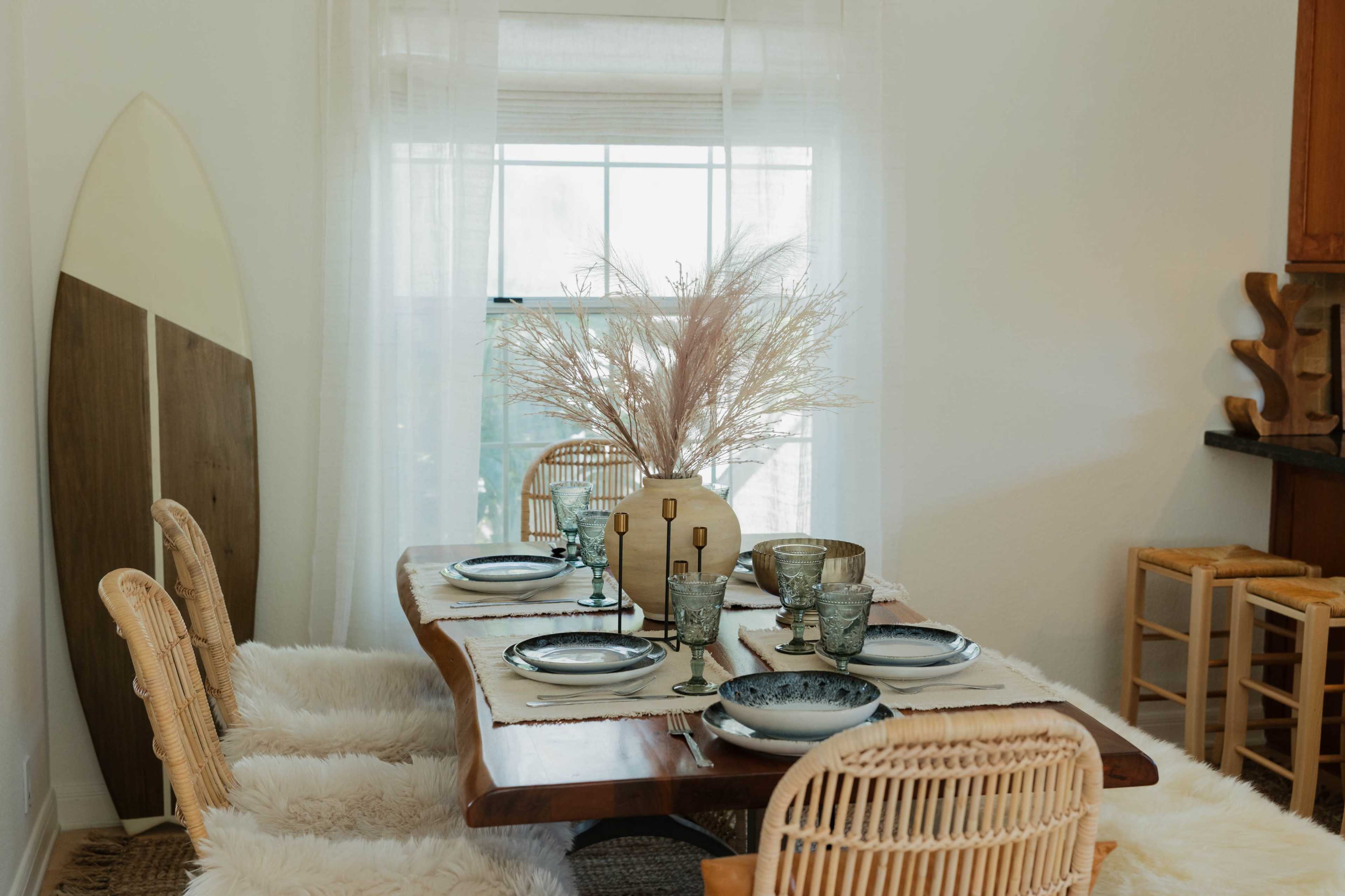 A dining table is set with plates, glasses, and a decorative centerpiece, accompanied by wicker chairs and a surfboard leaning against the wall.
