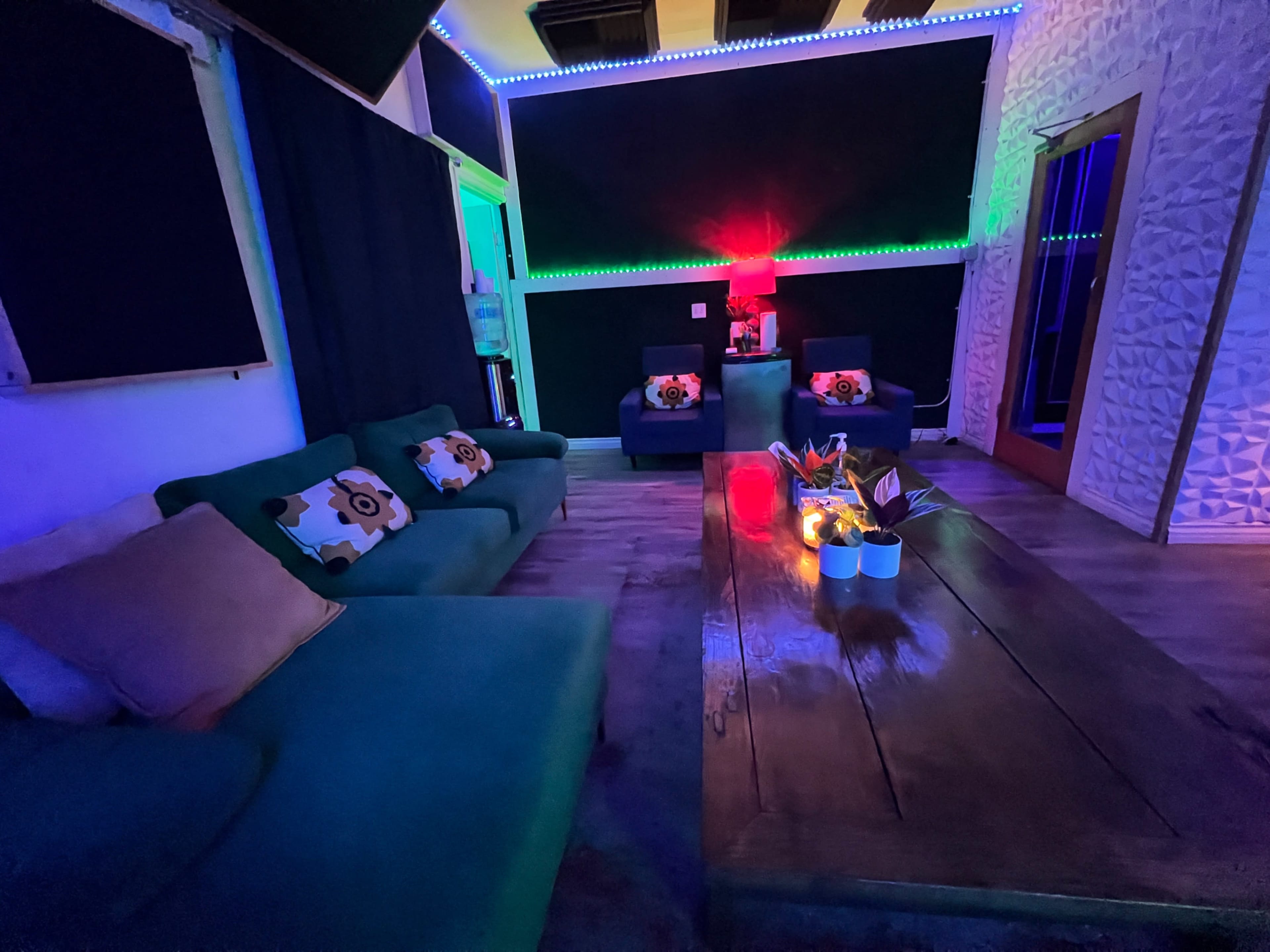 The image shows a dimly lit lounge area with colorful LED lights, featuring a large wooden table, two armchairs, and a green sofa adorned with floral-patterned cushions.