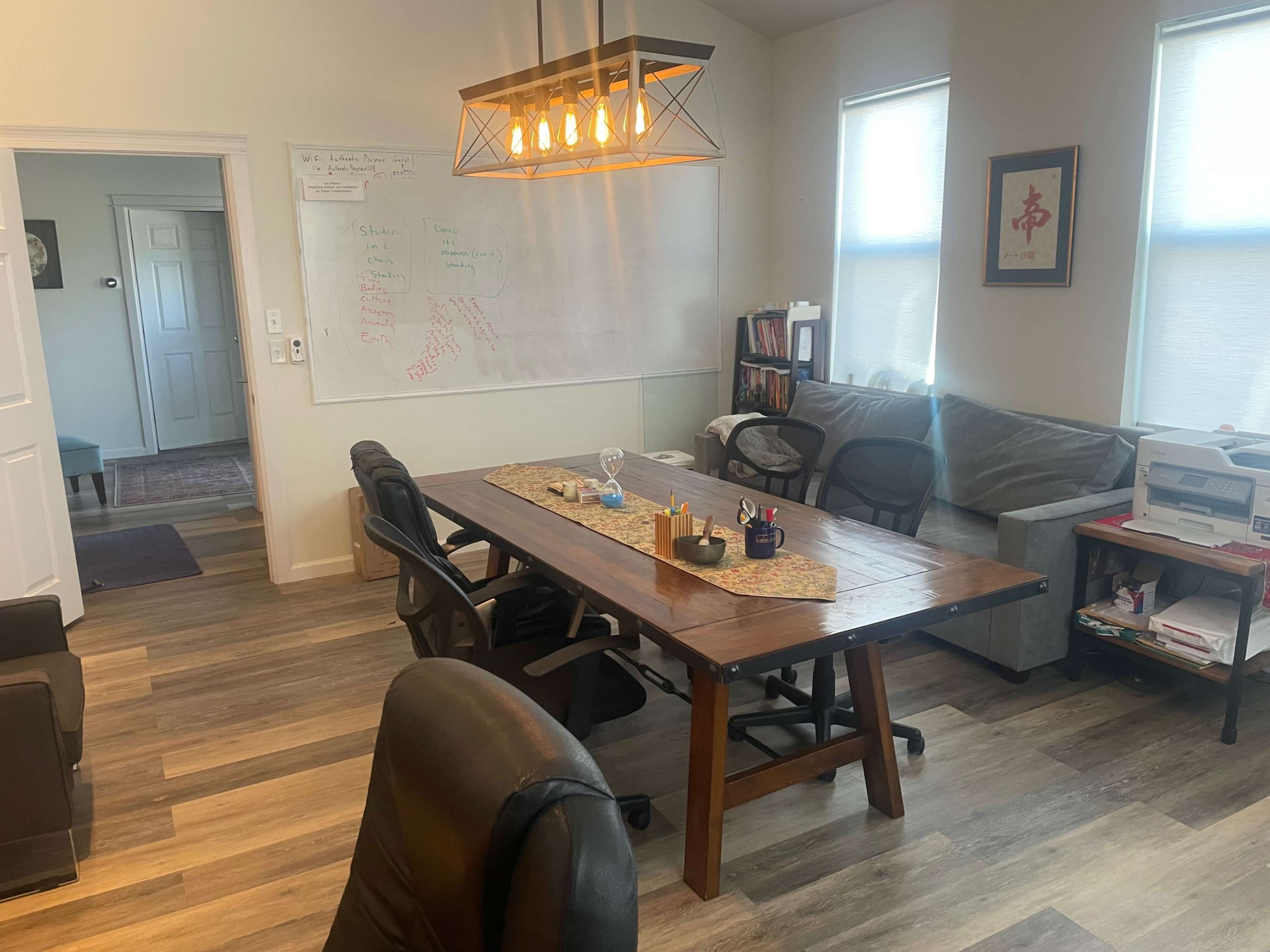 A brightly lit office space featuring a large wooden table with chairs, a whiteboard with notes, and bookshelves along the wall.