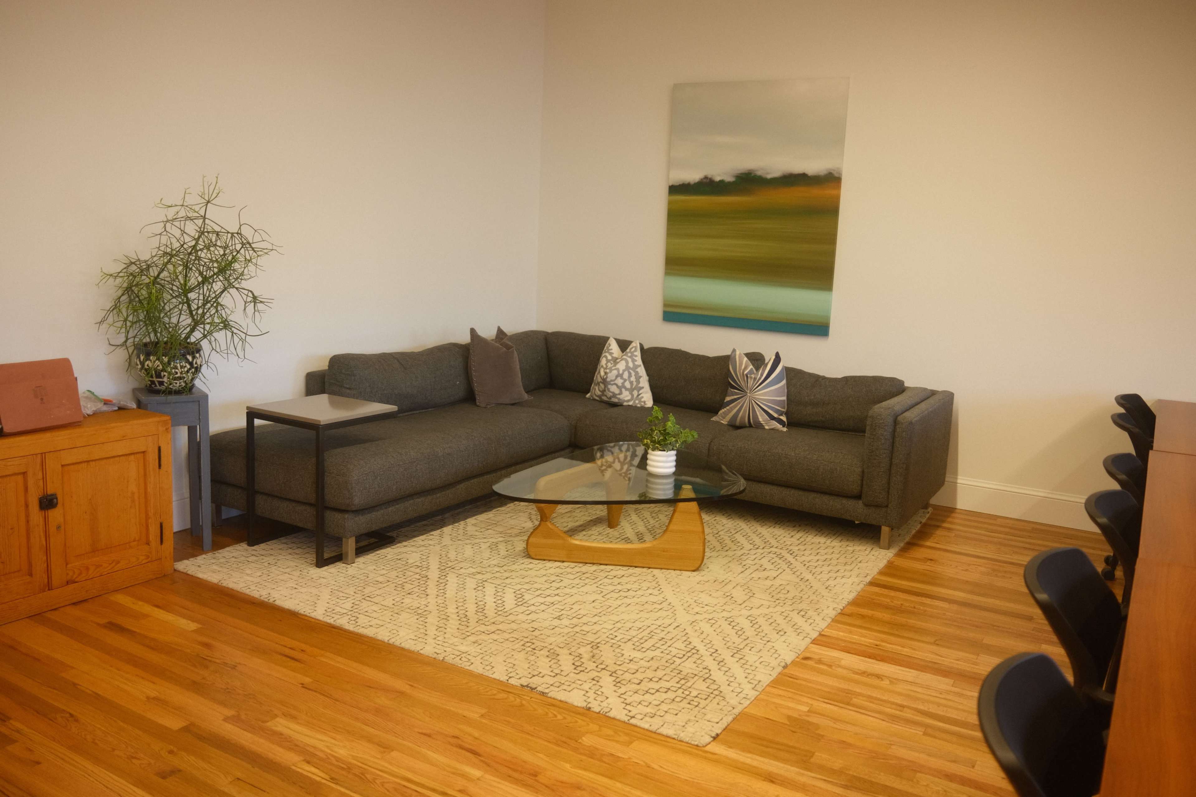 The image shows a cozy living room with a dark gray sectional sofa, a glass coffee table, a textured area rug, and a simple piece of artwork on the wall.
