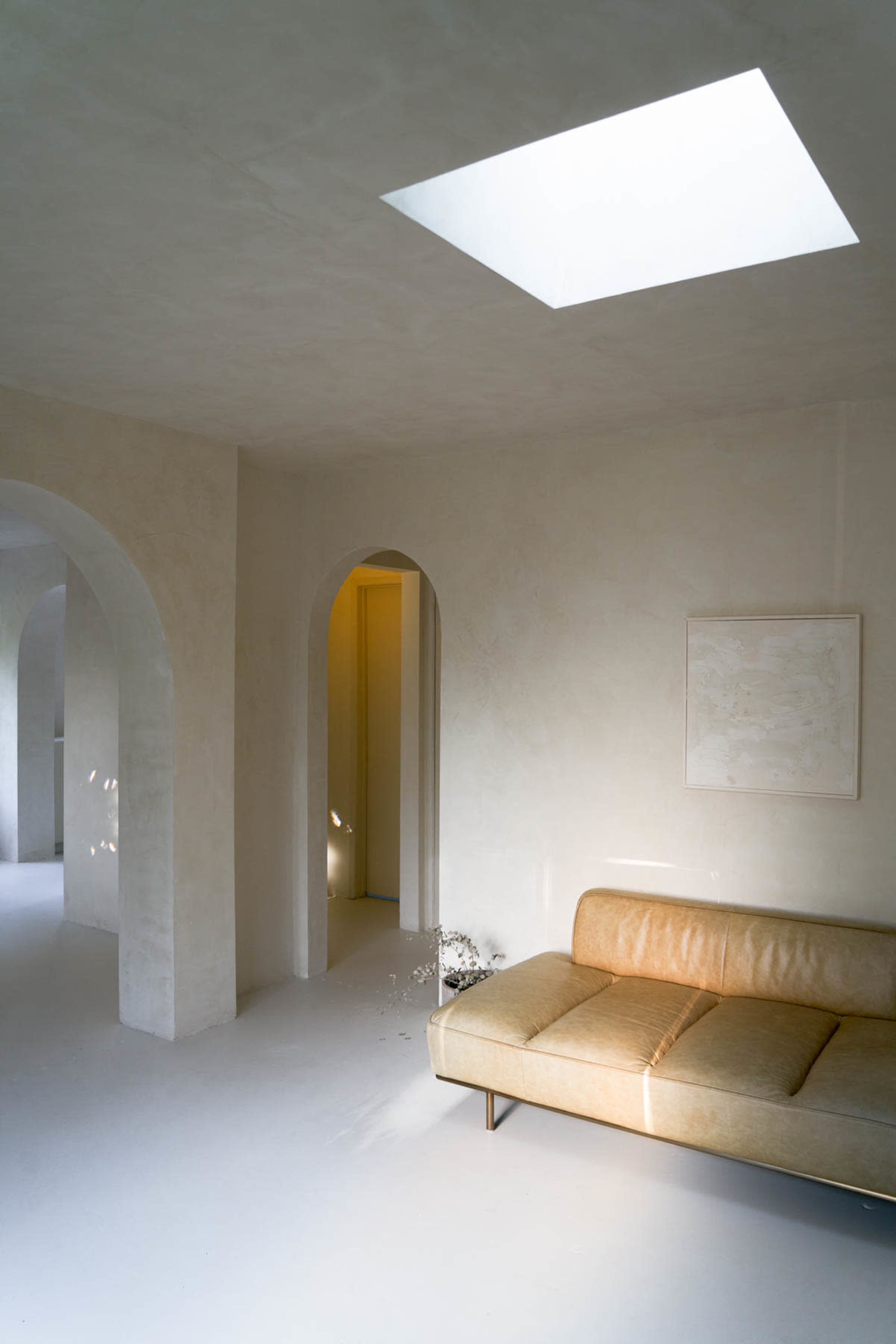 A minimalist interior features a beige sofa and arched doorways, illuminated by natural light from a skylight.