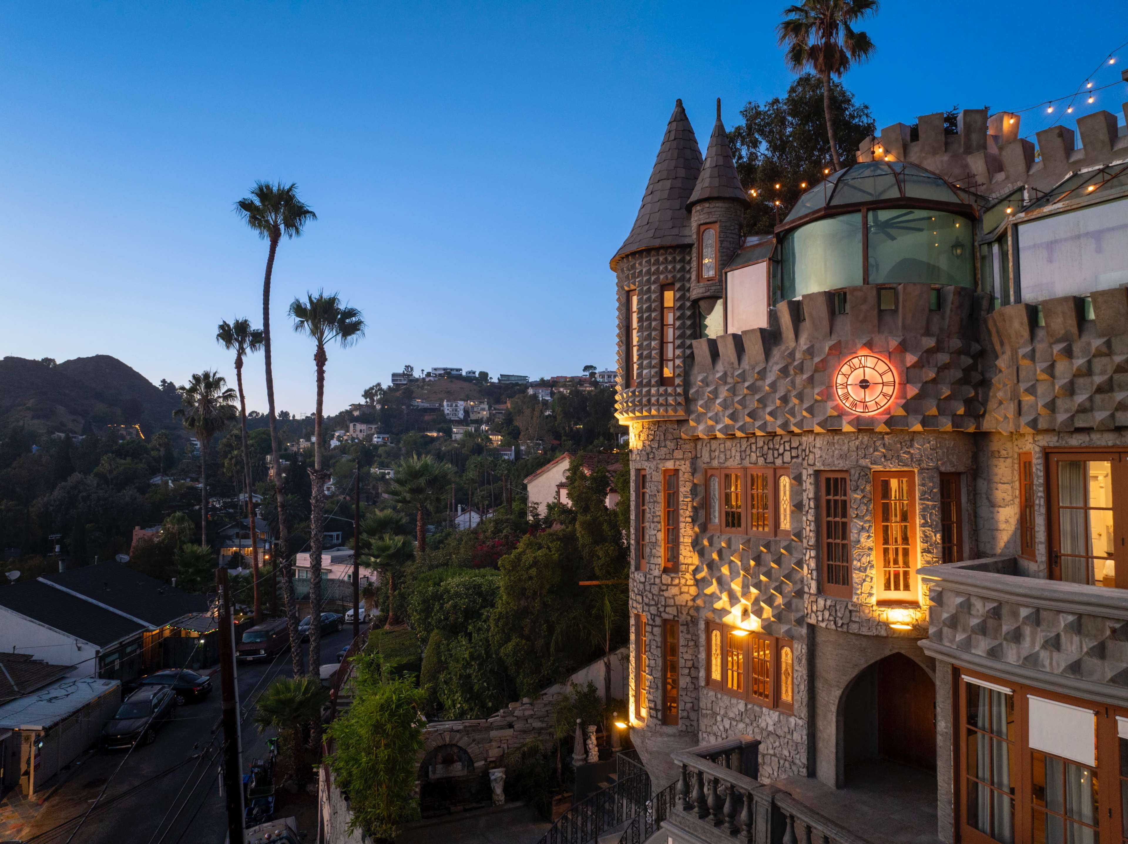The image shows a castle-like building with stone walls and turreted towers, illuminated by warm lights, set against a twilight sky with palm trees in the foreground.