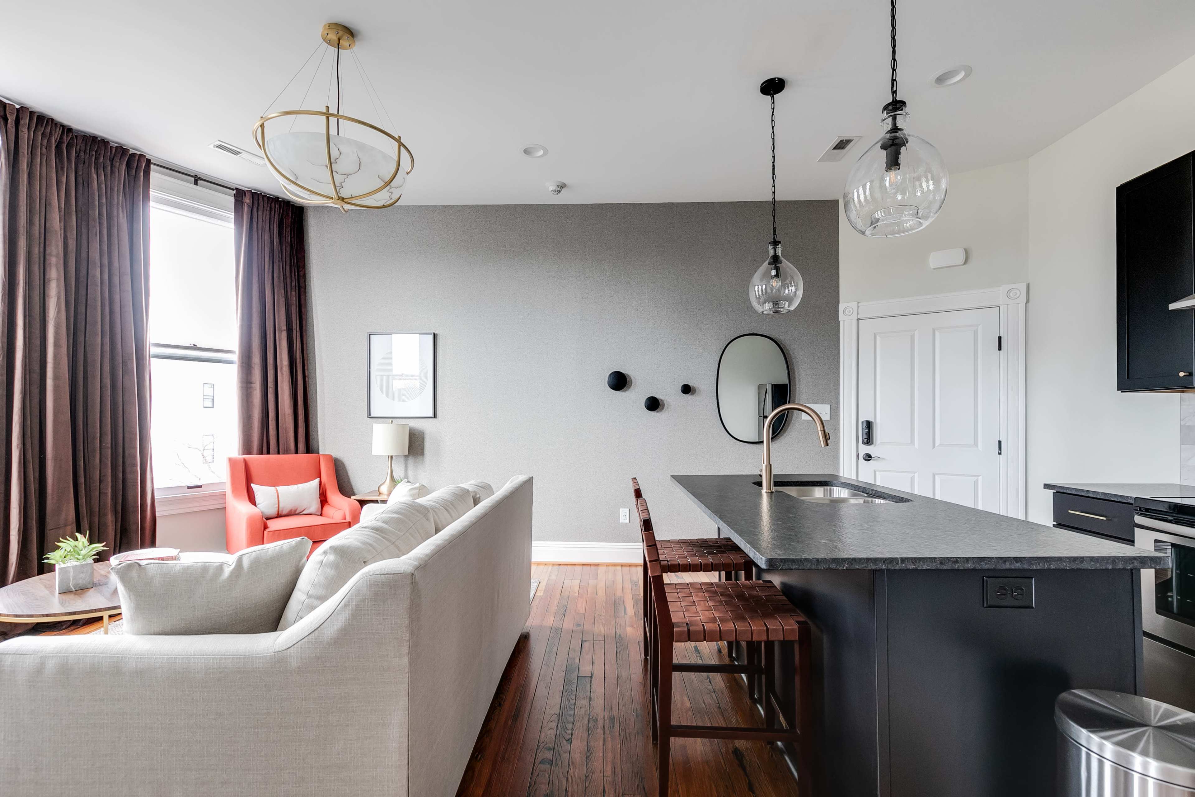 A modern living space features a sectional sofa, a kitchen island with bar stools, and large windows draped with dark curtains.