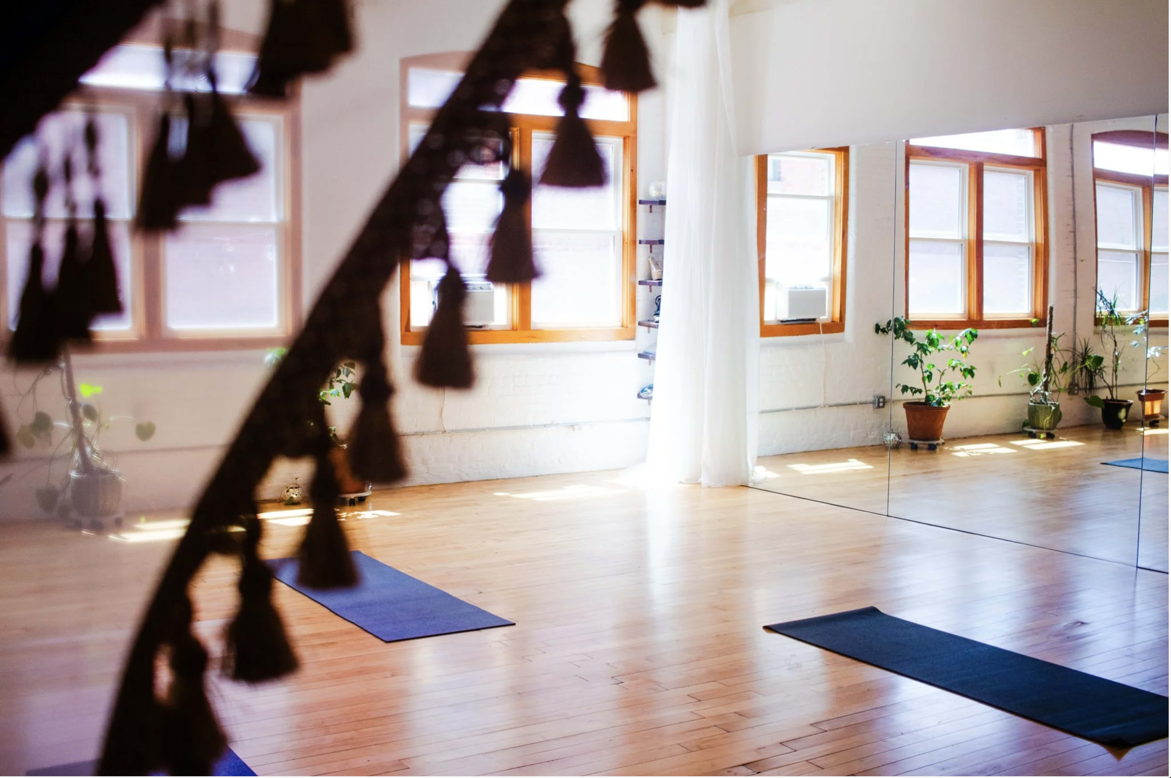The image shows a spacious yoga studio with wooden floors, large windows, and two yoga mats placed on the floor.