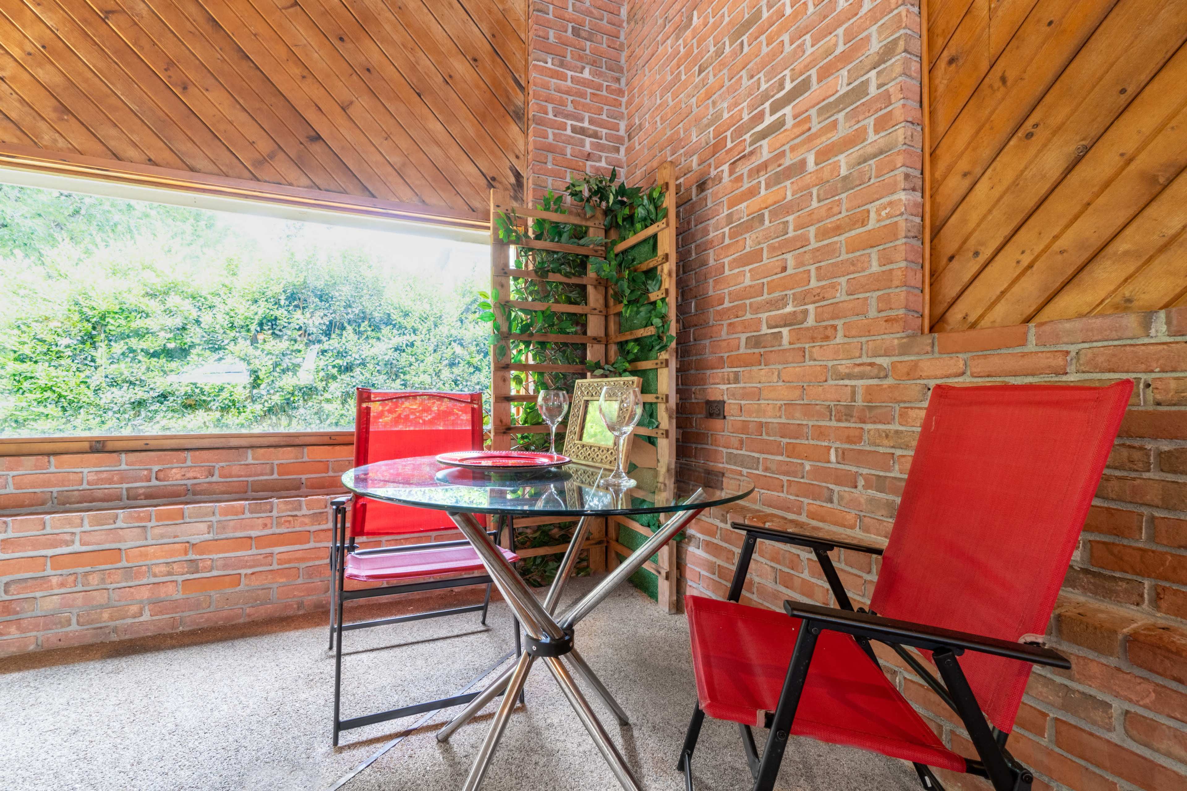 A small terrace features a glass table and two red chairs beside a wooden trellis with greenery.