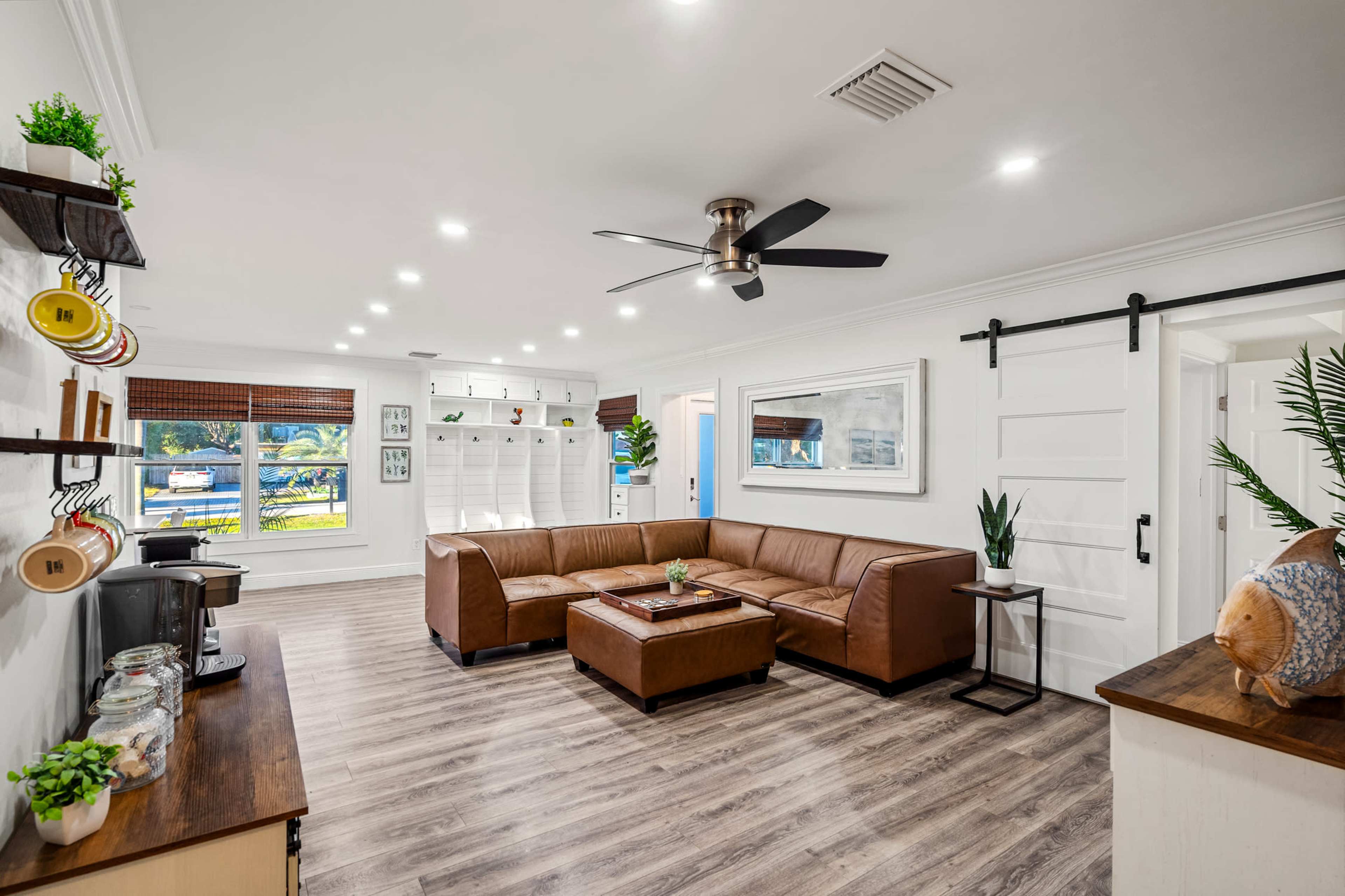 The living room features a brown sectional sofa, a wooden coffee table, and a ceiling fan, with bright lighting and a large window showcasing greenery outside.