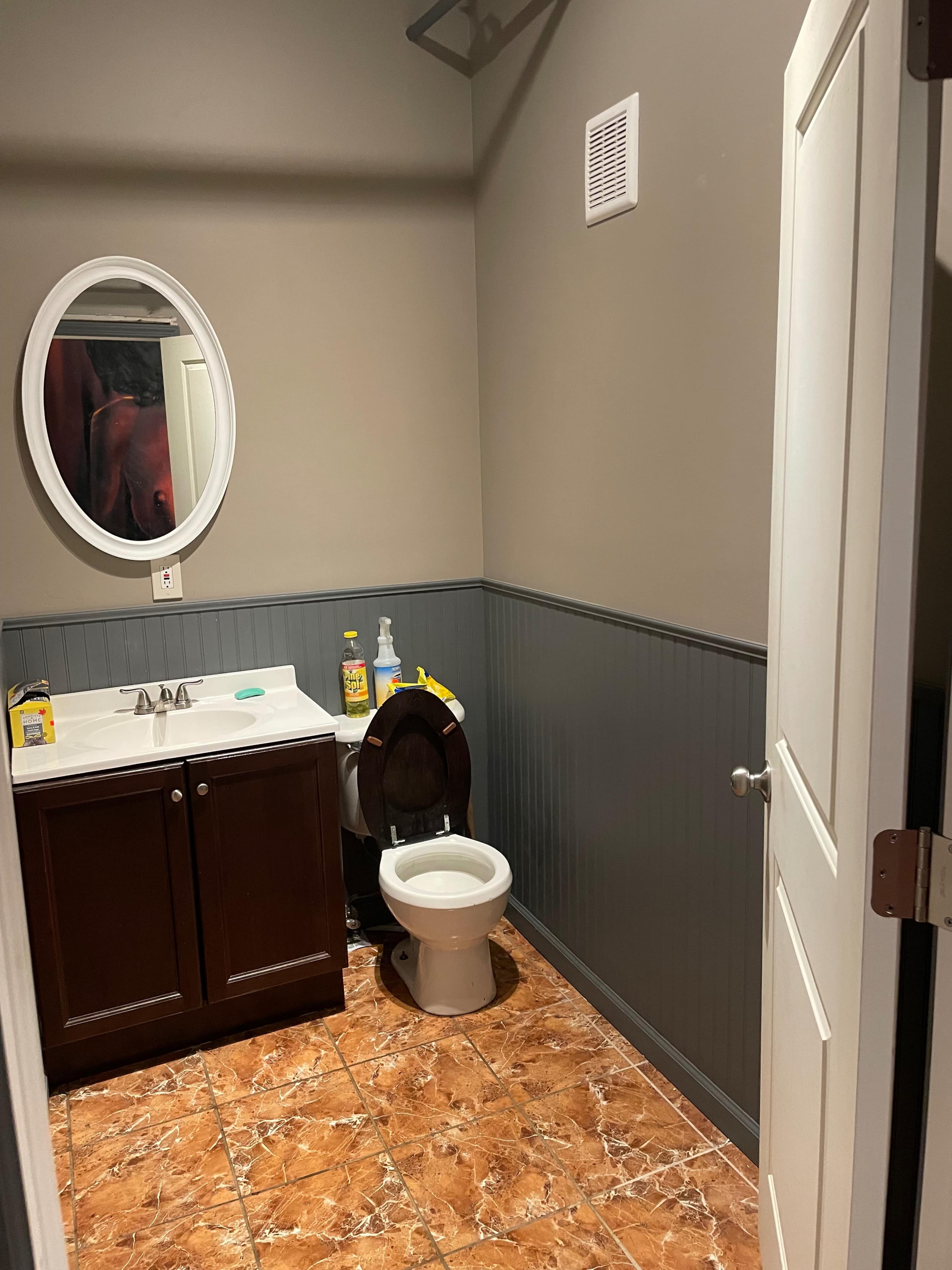 A small bathroom features a sink with cabinets, a toilet, and an oval mirror on the wall, with tiled flooring and gray wainscoting.